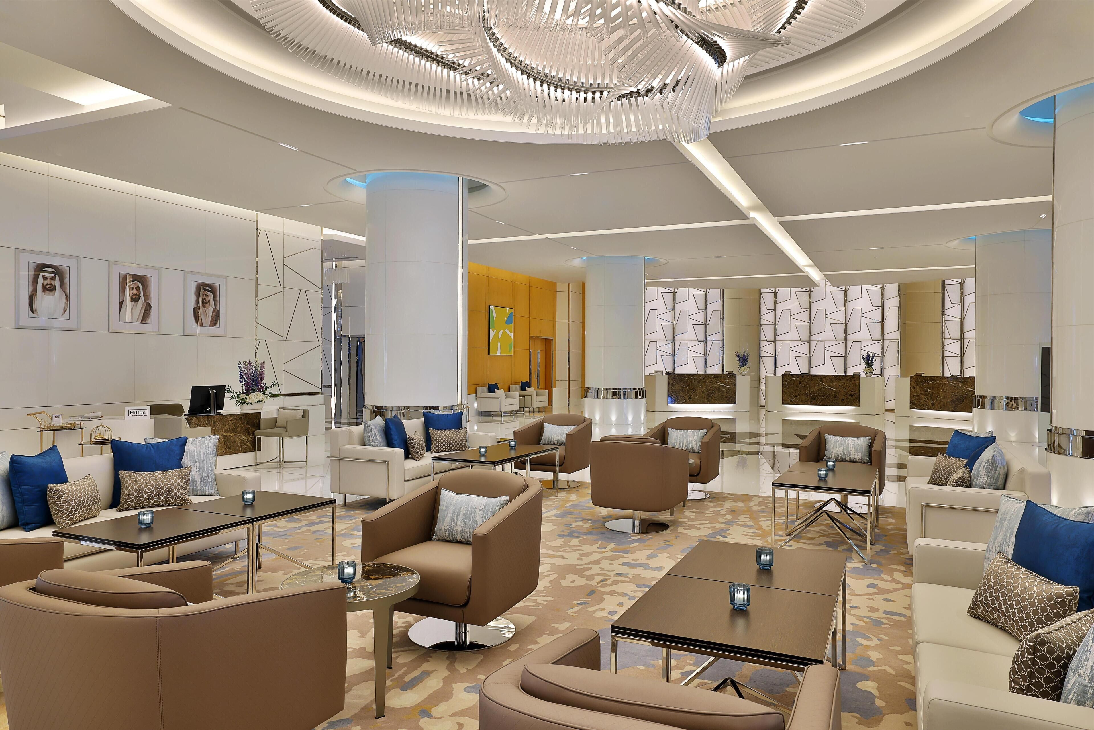 A modern hotel lobby with contemporary furniture, a central chandelier, and a reception area.
