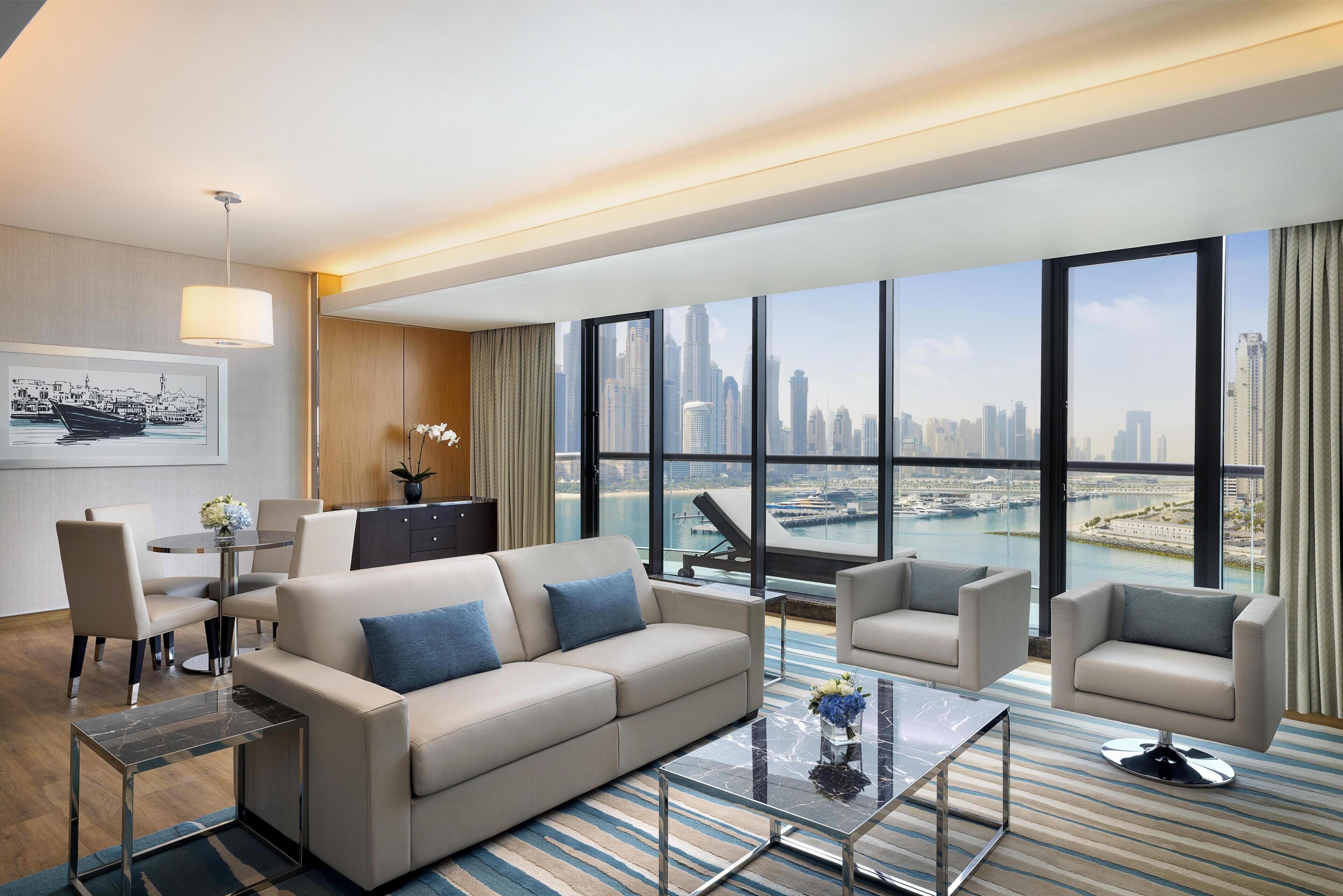 A modern hotel room with a large window offering a city view, a sofa, armchairs, a coffee table, and a dining area with chairs and a table.