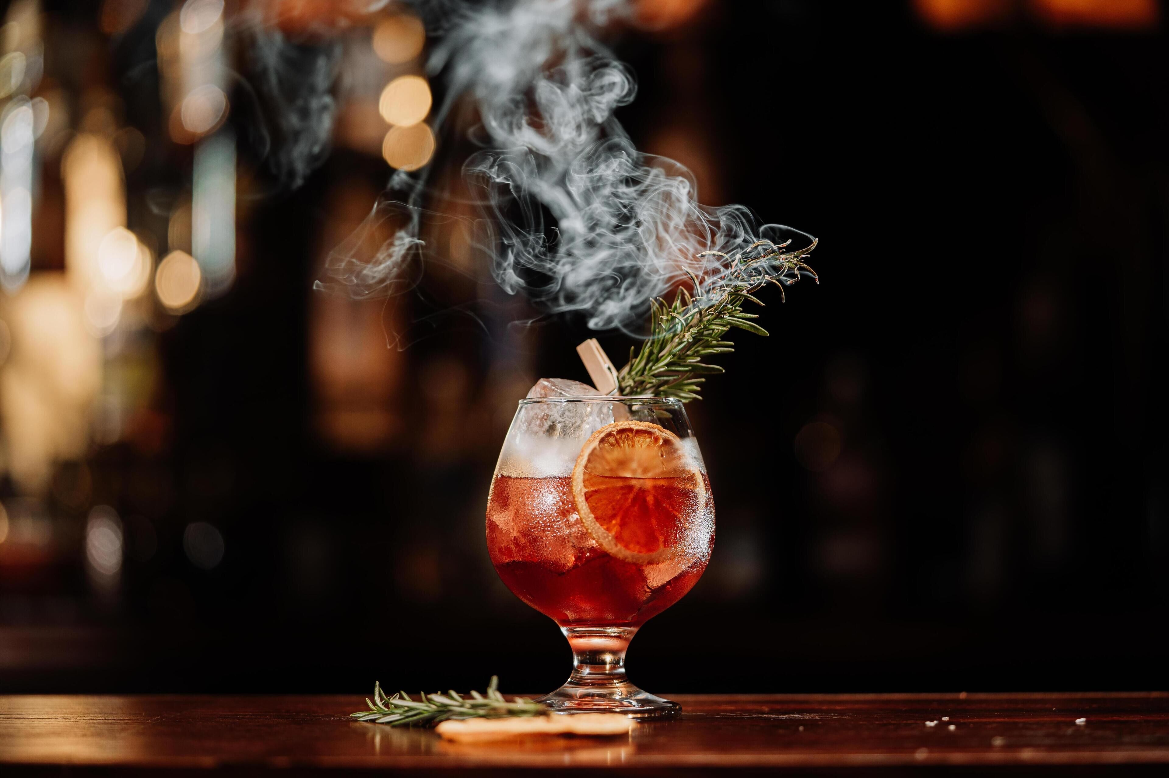 Een cocktail met ijs, een oranje schijfje en een takje rozemarijn op een bargedeelte.