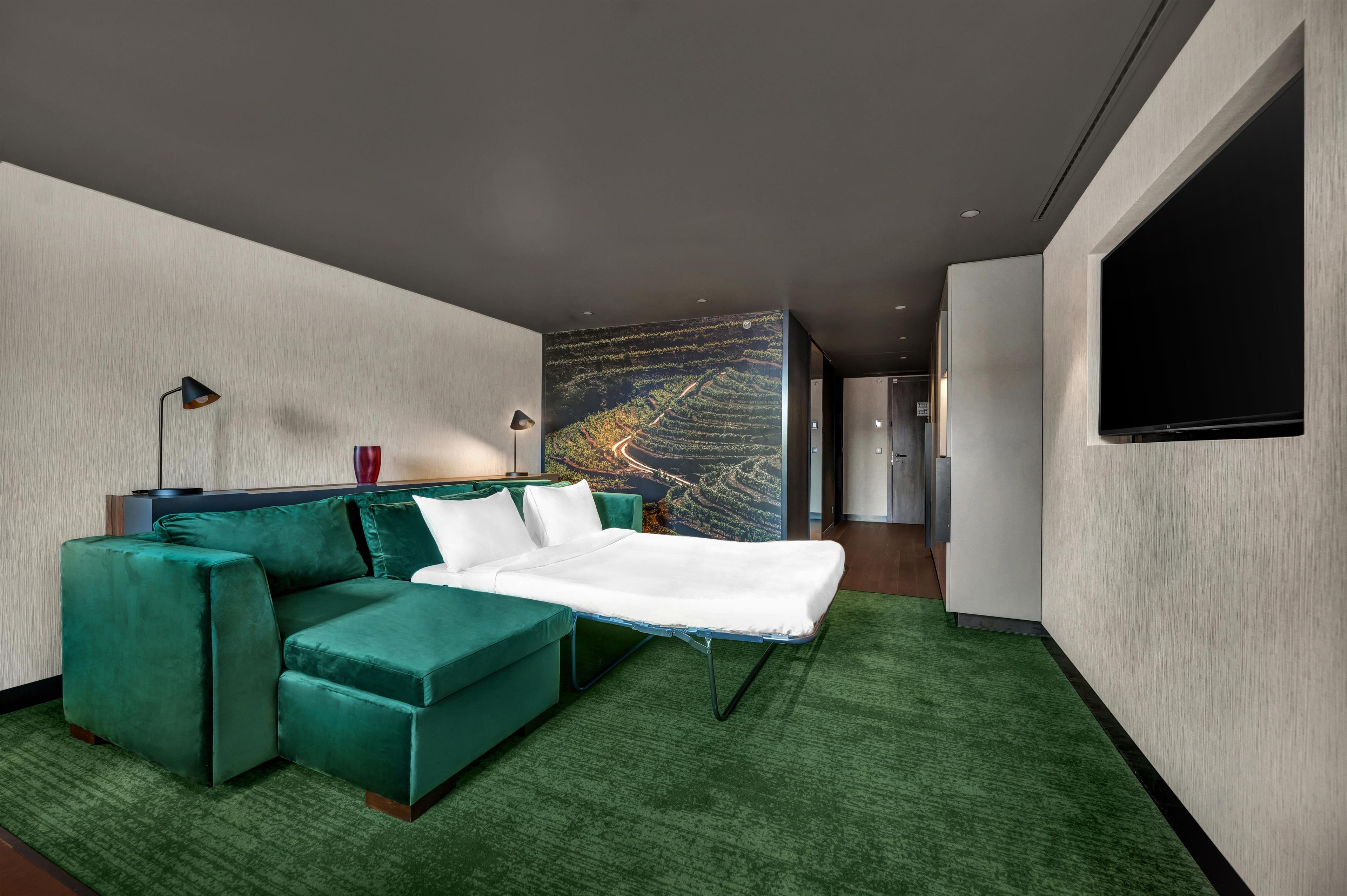 A modern hotel room with a green sofa, a flat-screen TV, and a large window with a scenic view.