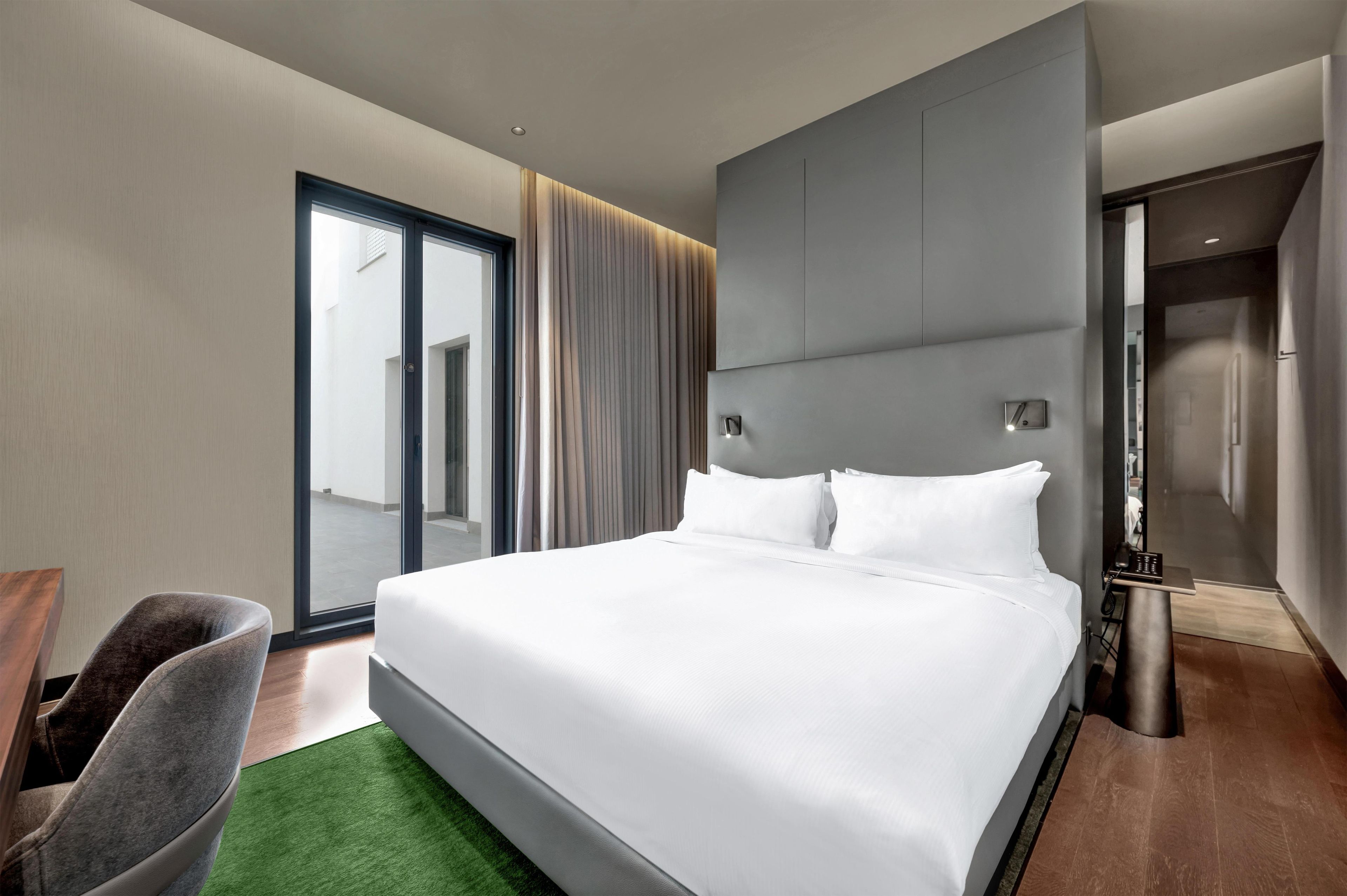 Une chambre d’hôtel moderne dotée d’un grand lit, d’un coin salon avec un fauteuil et d’une vue sur l’extérieur, visible à travers une porte vitrée.
