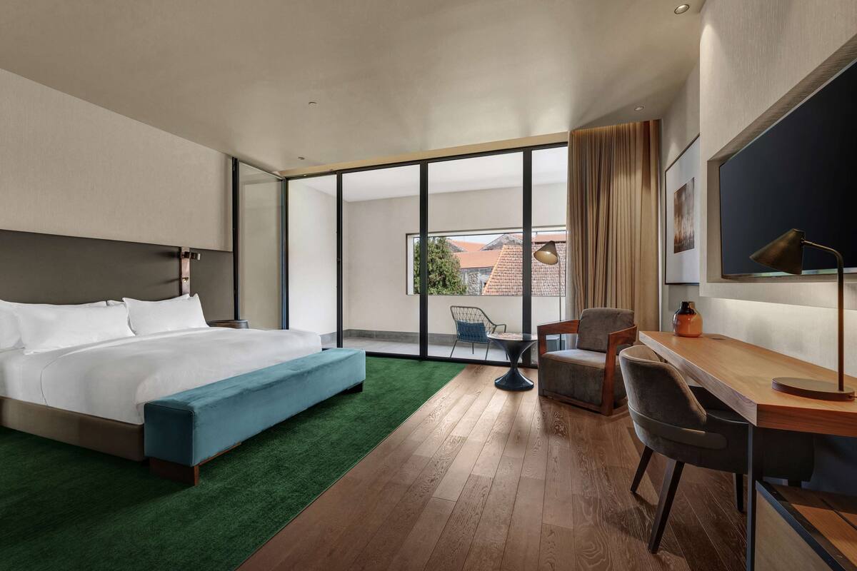 A modern hotel room with a large bed, a desk, a chair, and a view of the outdoors.