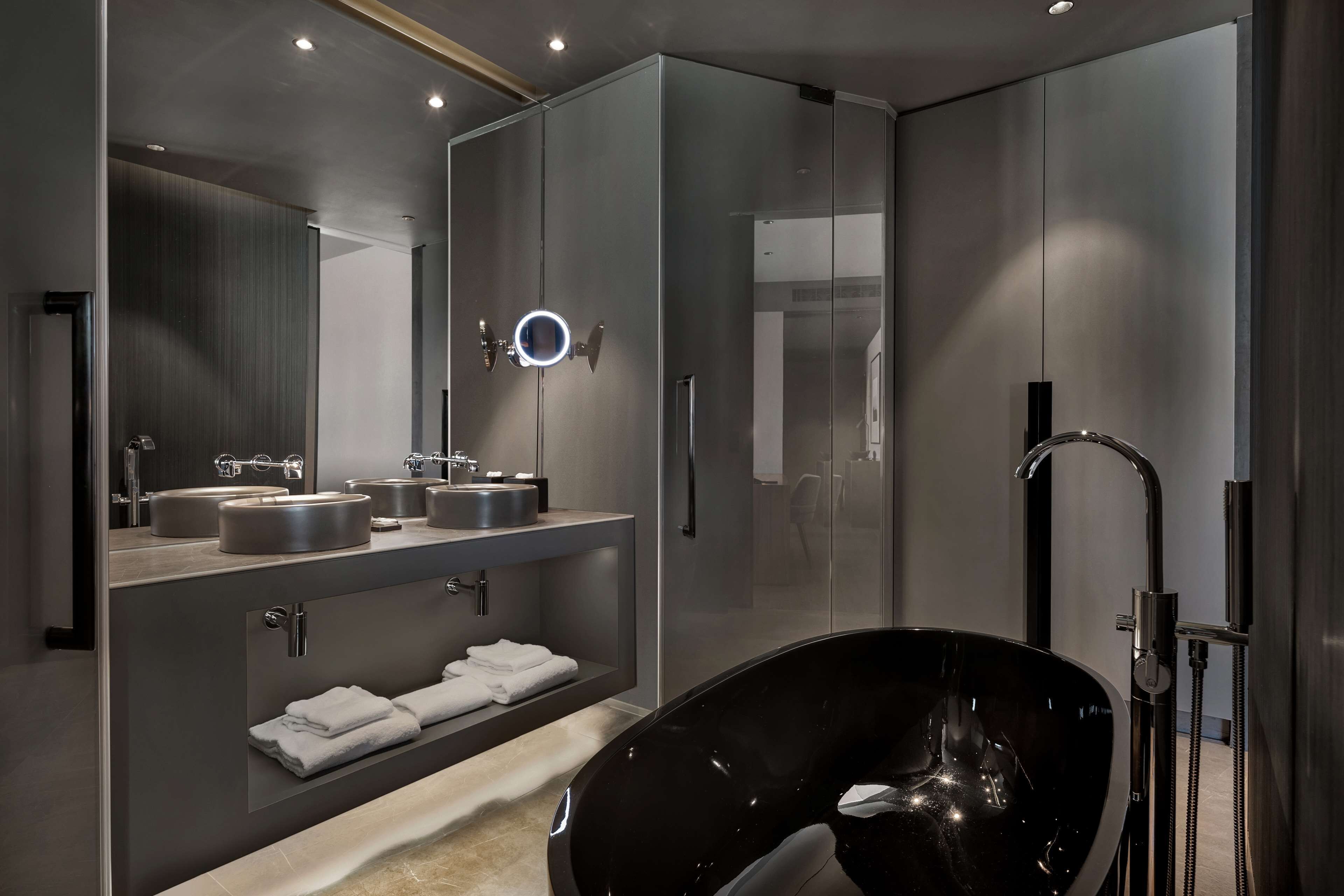 A modern bathroom with a large black bathtub, a round mirror with lights, and a sleek vanity with multiple sinks.