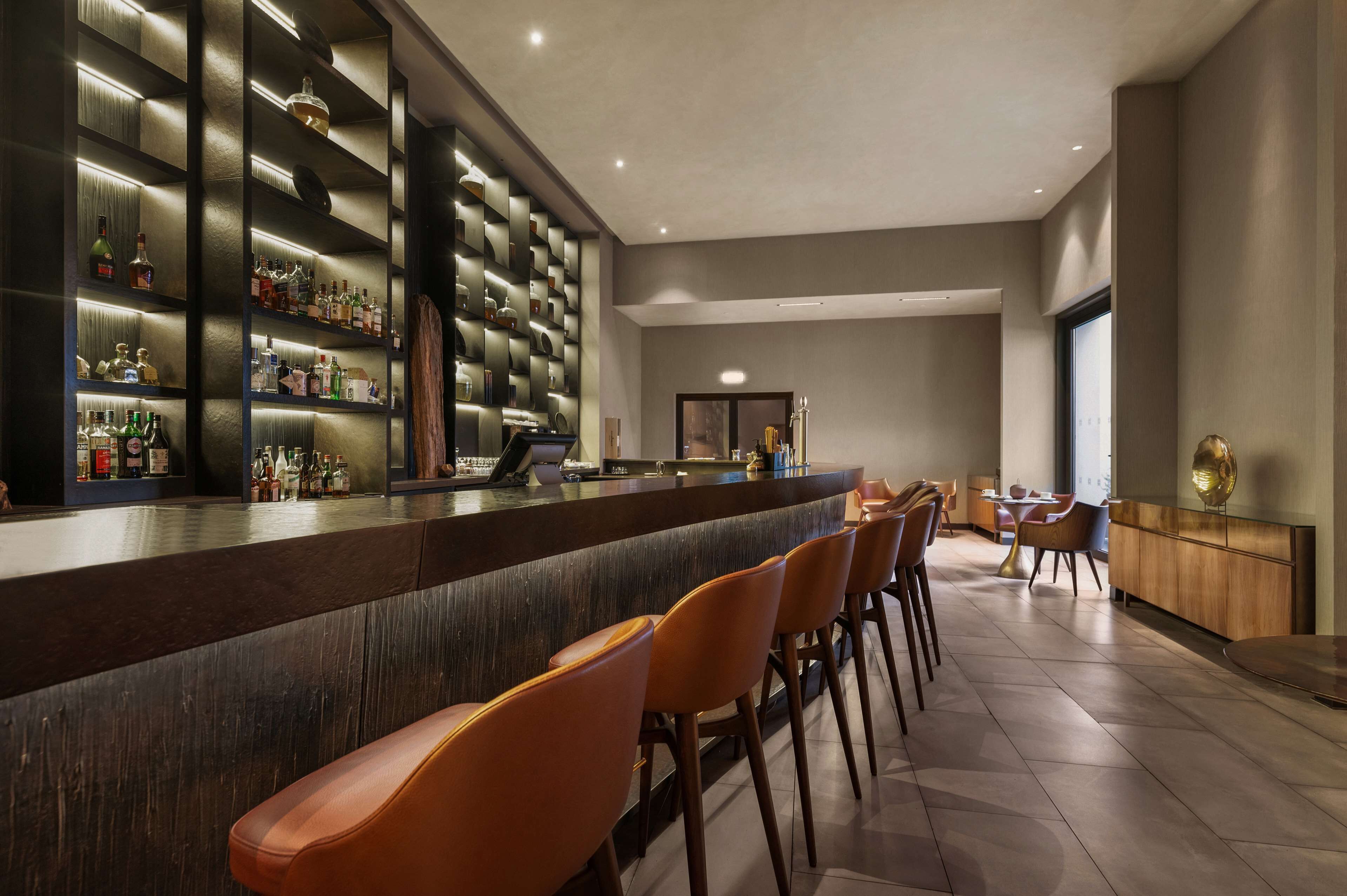 Uma área de bar com um balcão comprido, várias prateleiras com garrafas e algumas cadeiras laranja.