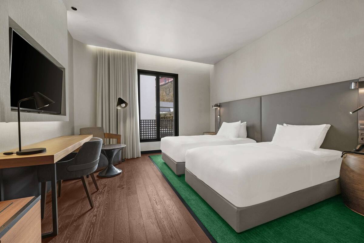A hotel room with two beds, a desk with a chair, a TV, and a large window with curtains.