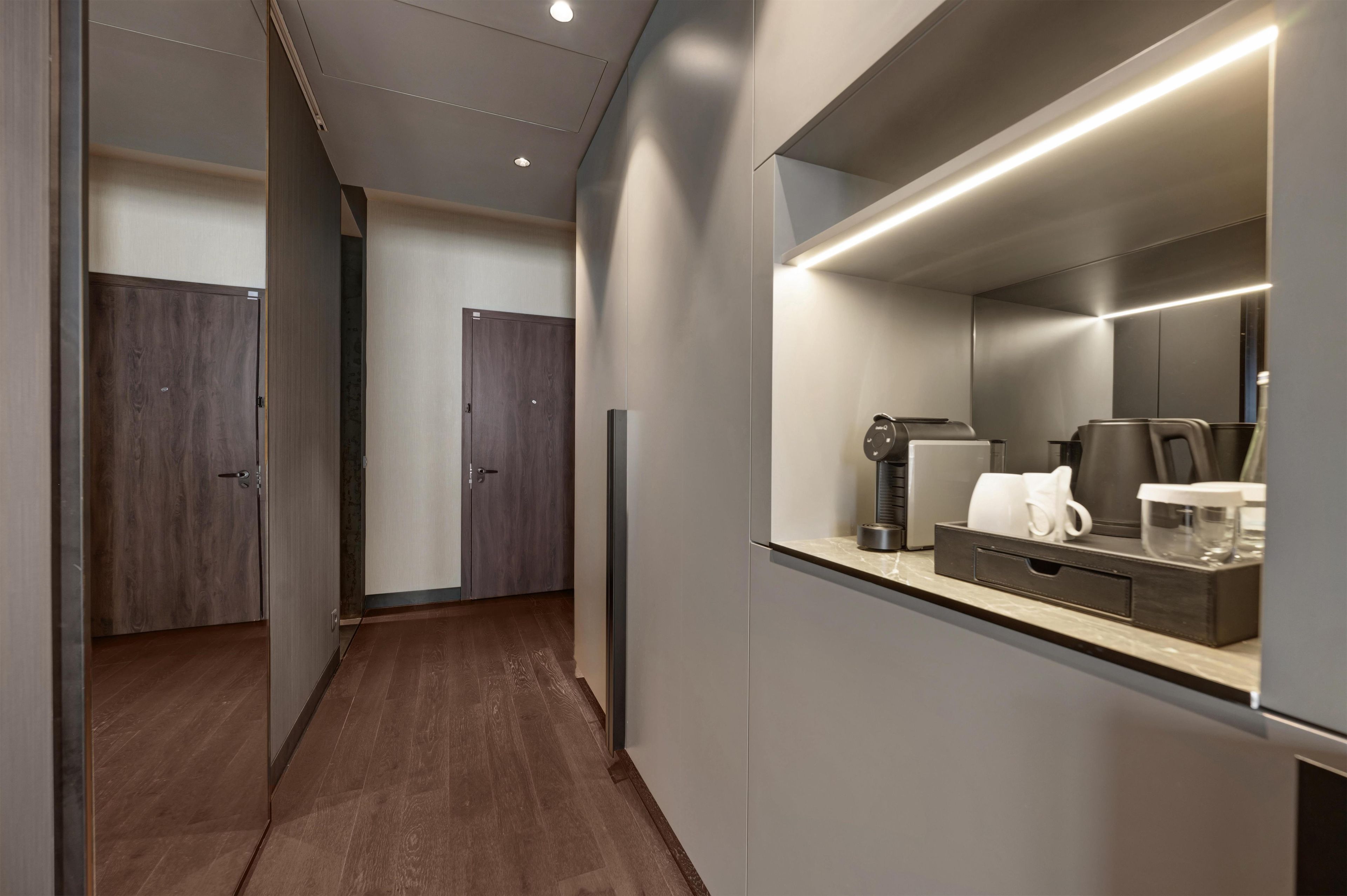 A modern hotel room with a wooden floor, a large mirror, a coffee machine, and a small table with cups and a kettle.