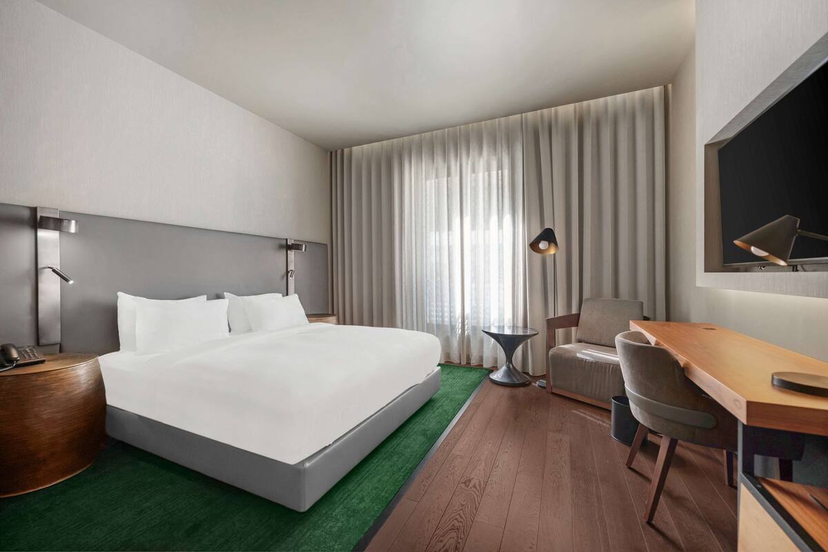 A modern hotel room with a large bed, a desk, a chair, a small table, and a TV mounted on the wall.