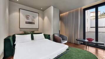 Quarto de hotel moderno com cama, cadeira, mesa pequena e grande janela com cortinas.