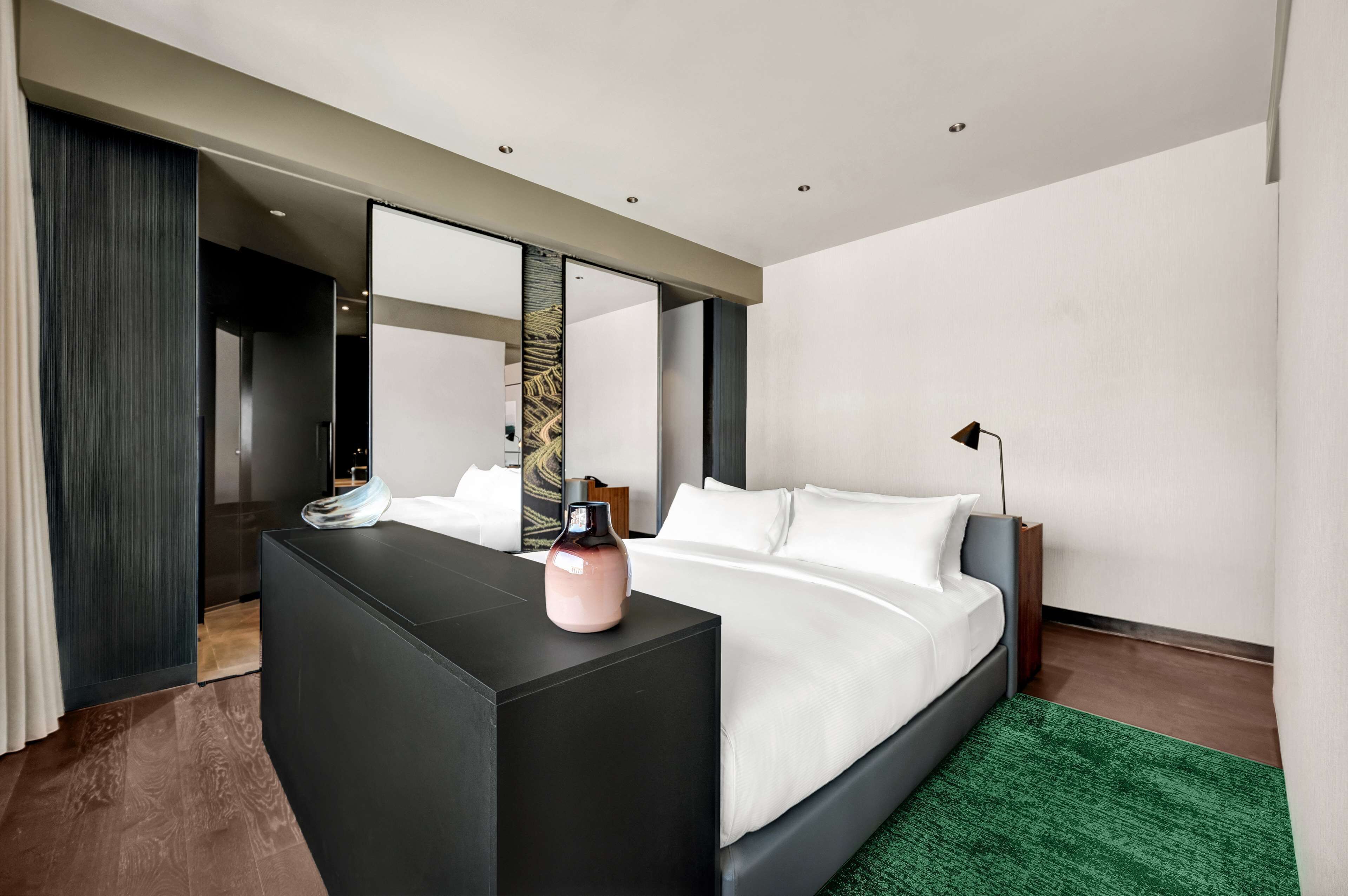 Ein modernes Hotelzimmer mit einem großen Bett, einer dunkelholzigen Kommode, einem grünen Teppich und einem großen Spiegel, der den Raum reflektiert.
