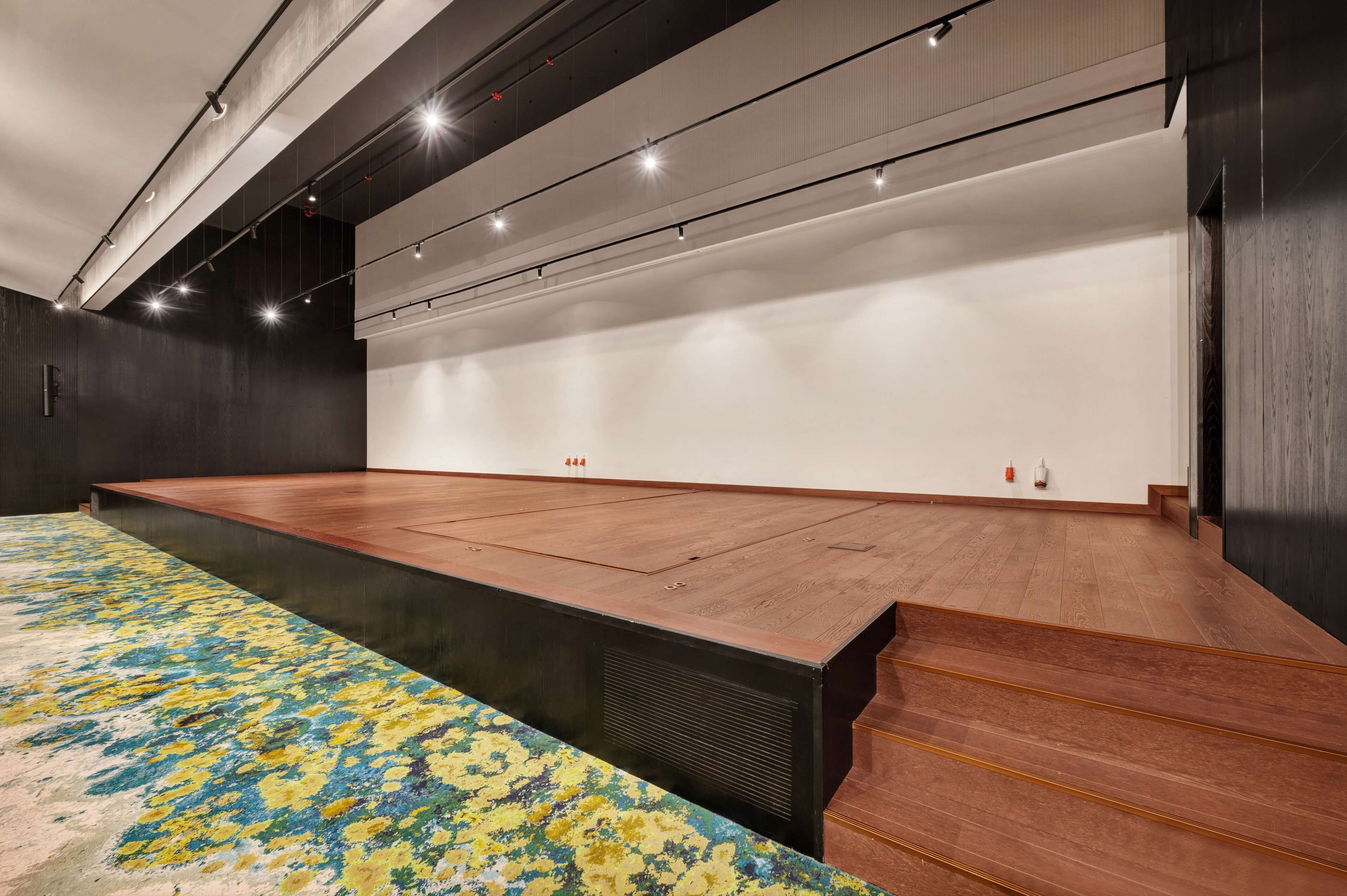 A spacious room with a large wooden stage, a patterned carpet, and a high ceiling with track lighting.