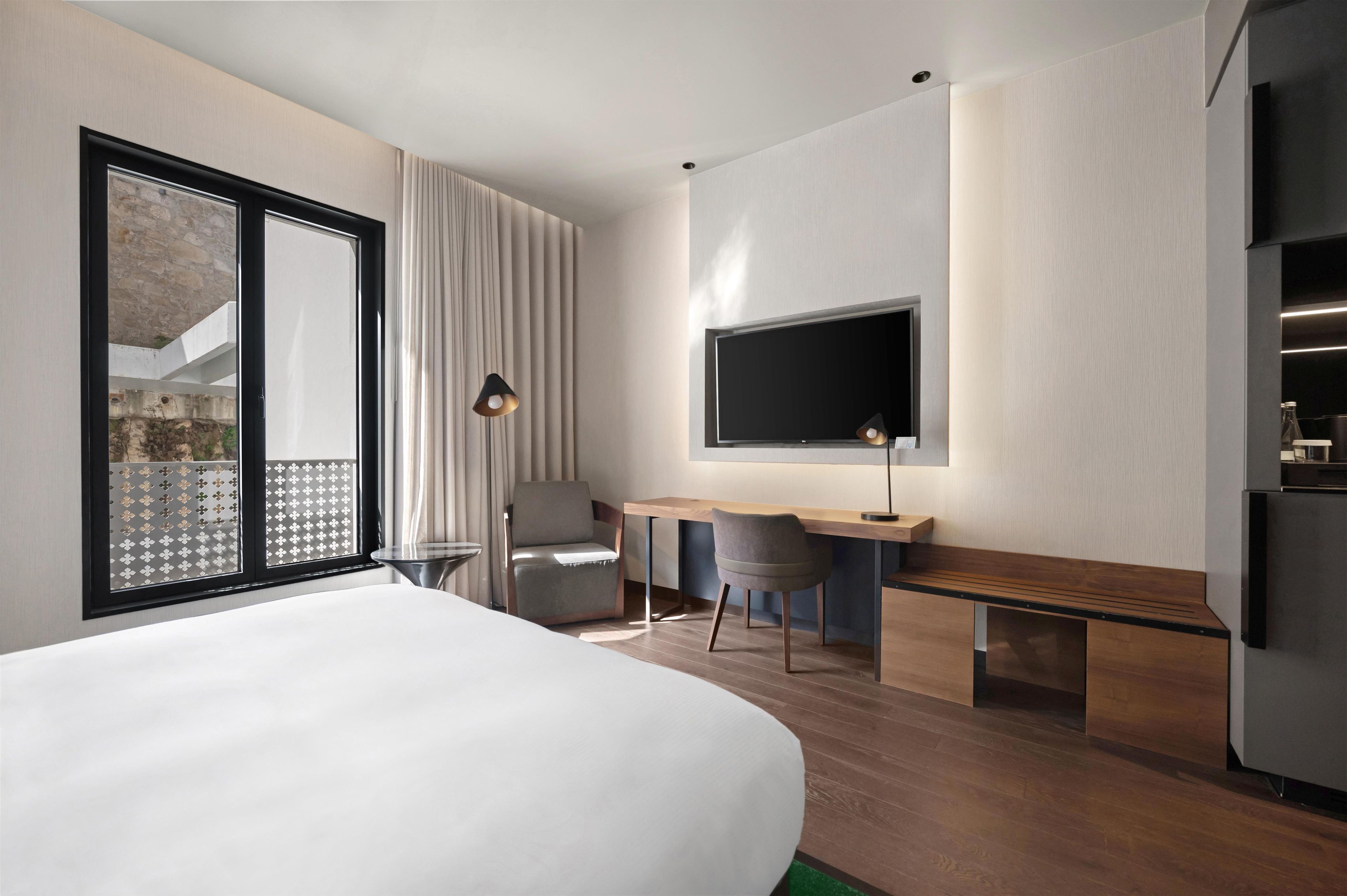 A modern hotel room with a large bed, a desk, a chair, a TV, and a refrigerator.