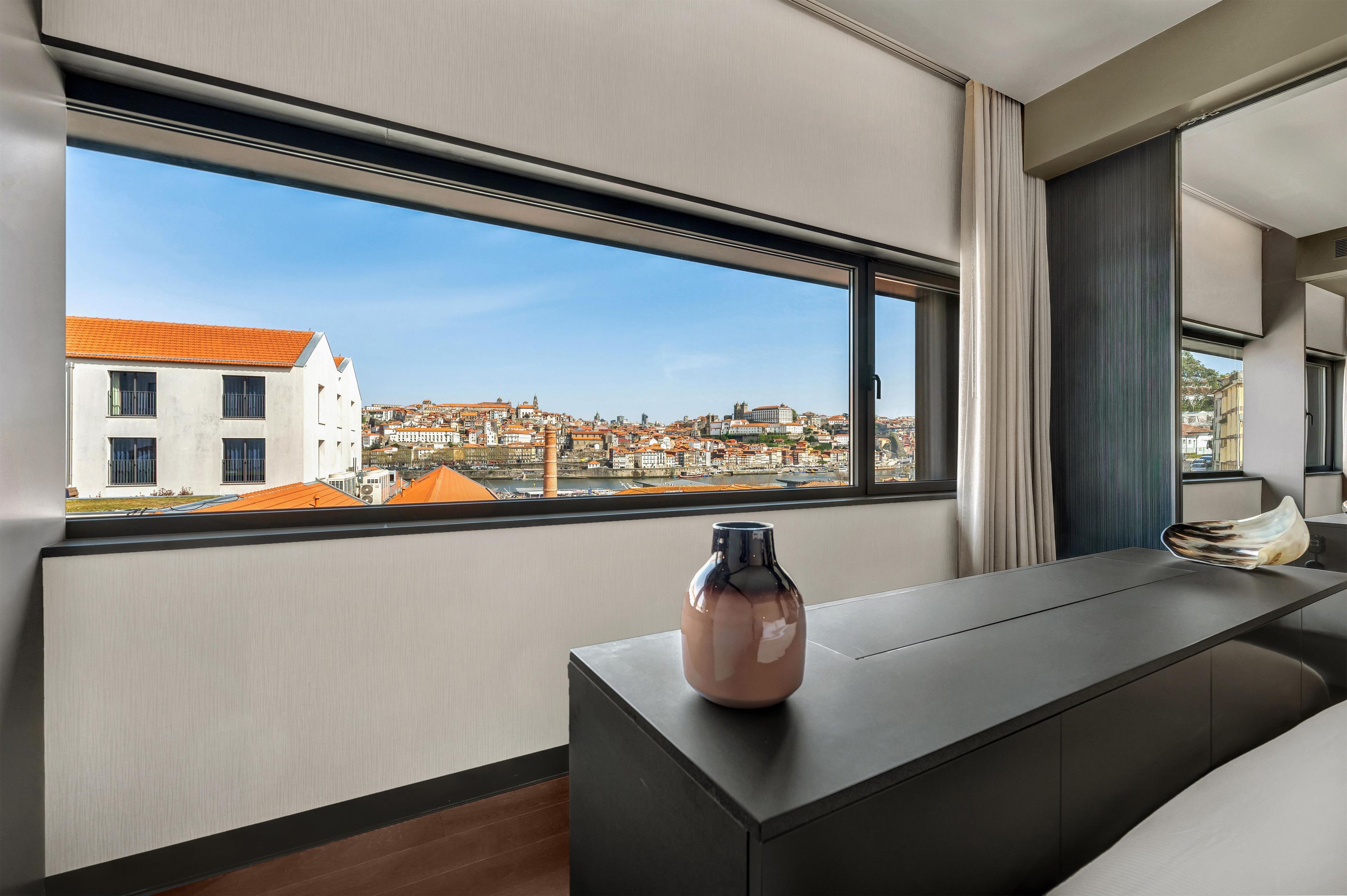 A modern hotel room with a large window offering a city view, a dark wooden desk, and a vase on the table.