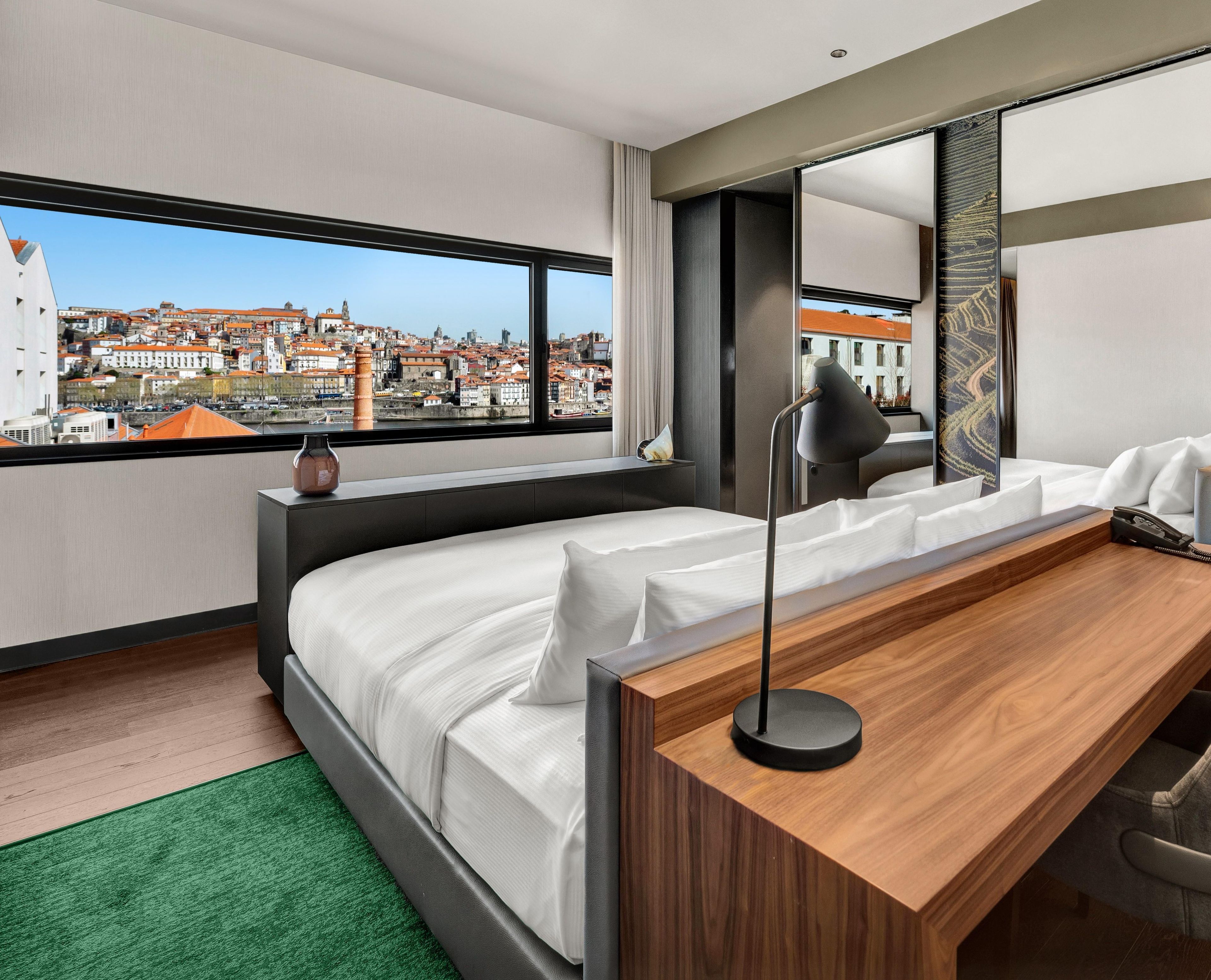 Moderni hotellihuone, jossa on suuri sänky, puinen työpöytä ja näkymä kaupunkimaisemaan.