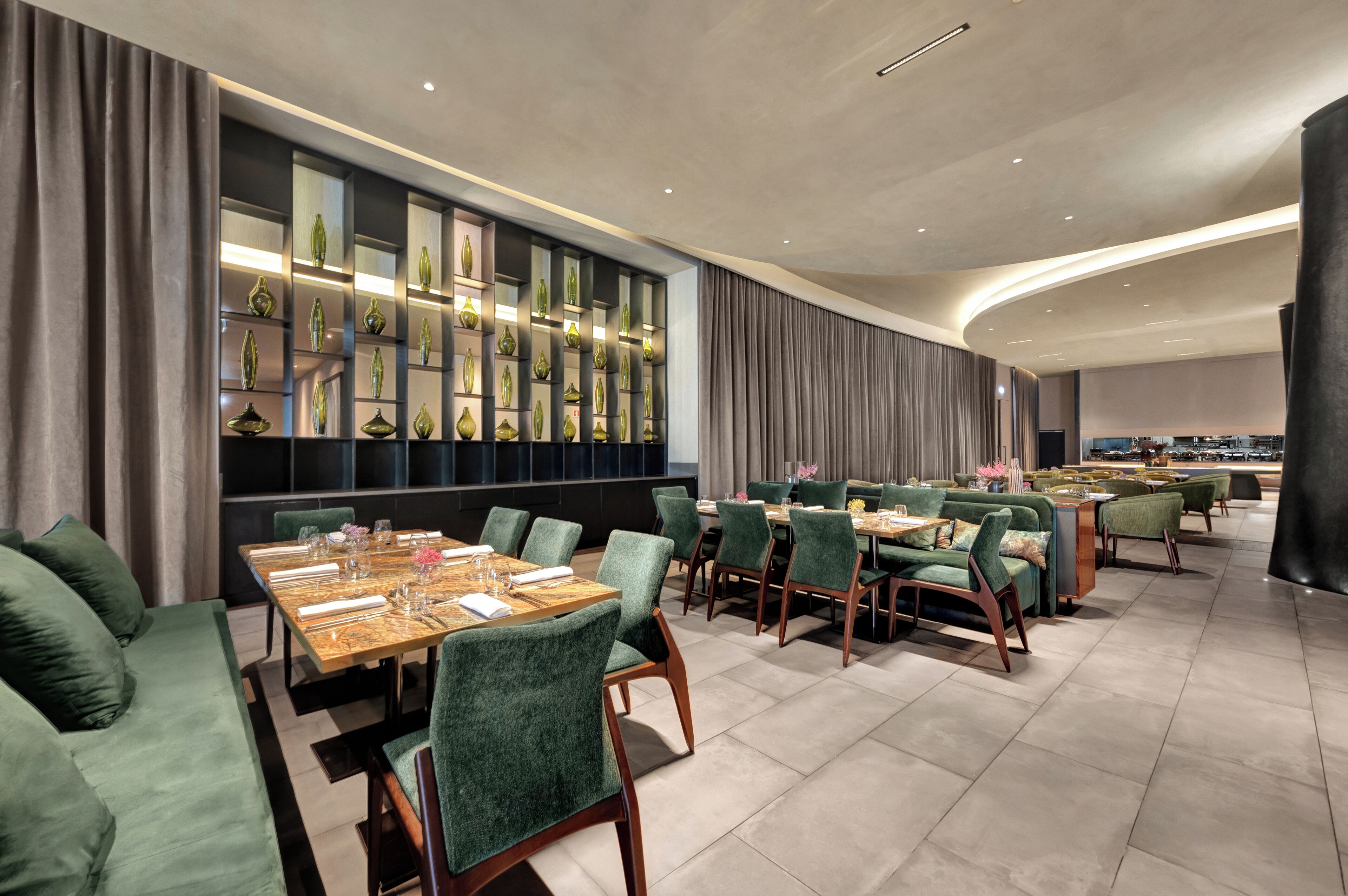 A modern dining area with green upholstered chairs, a wooden table set for a meal, and a wall of decorative vases.