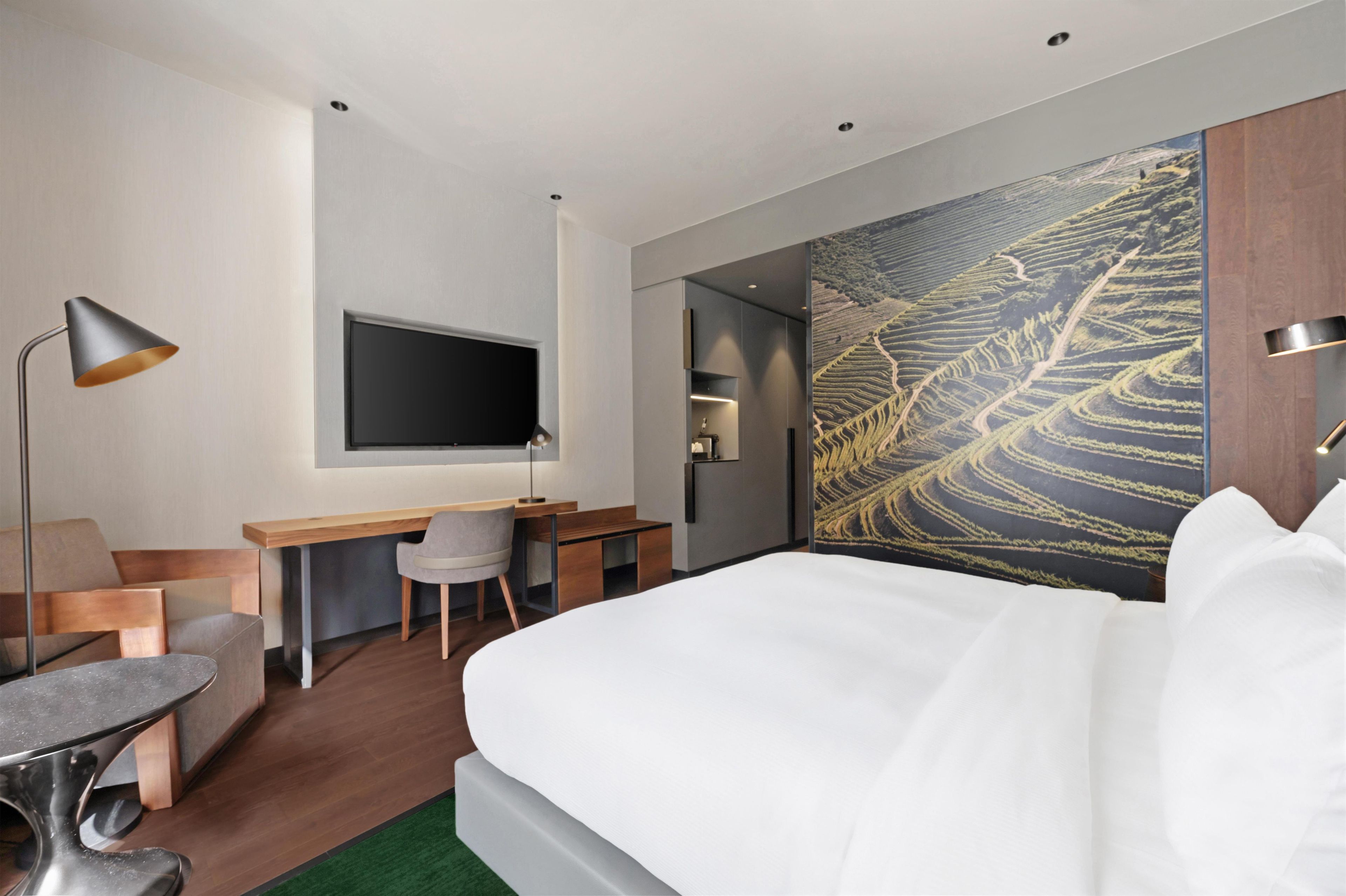 Et moderne hotelværelse med seng, skrivebord, stol og et stort TV på væggen.