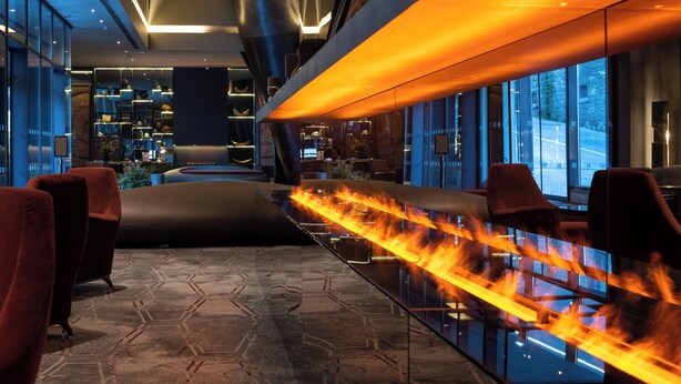 A modern hotel lobby with a glass partition, seating area, and a fire feature.