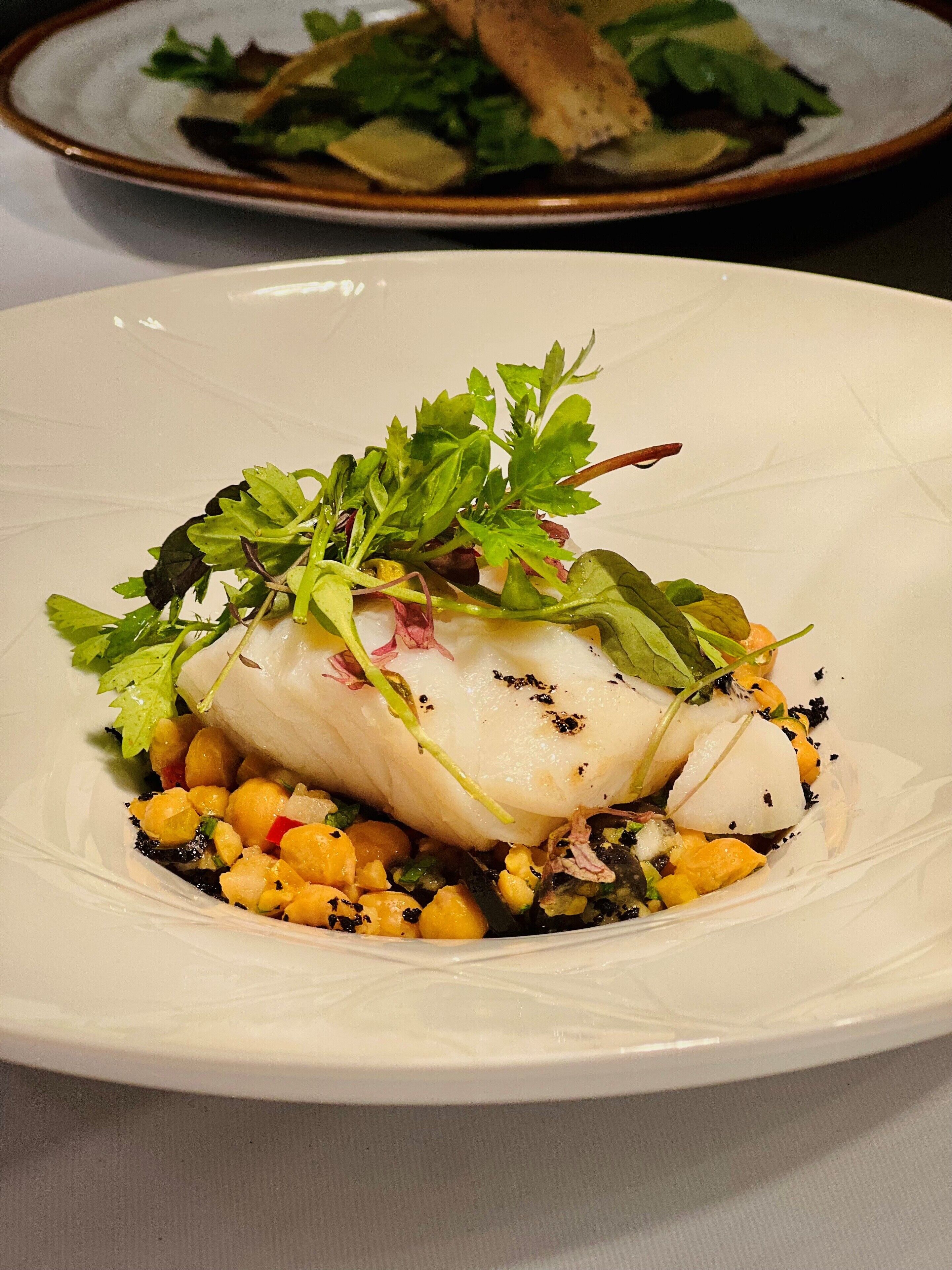 Um prato branco com um ensopado de peixe, adornado com folhas verdes e grão-de-bico.