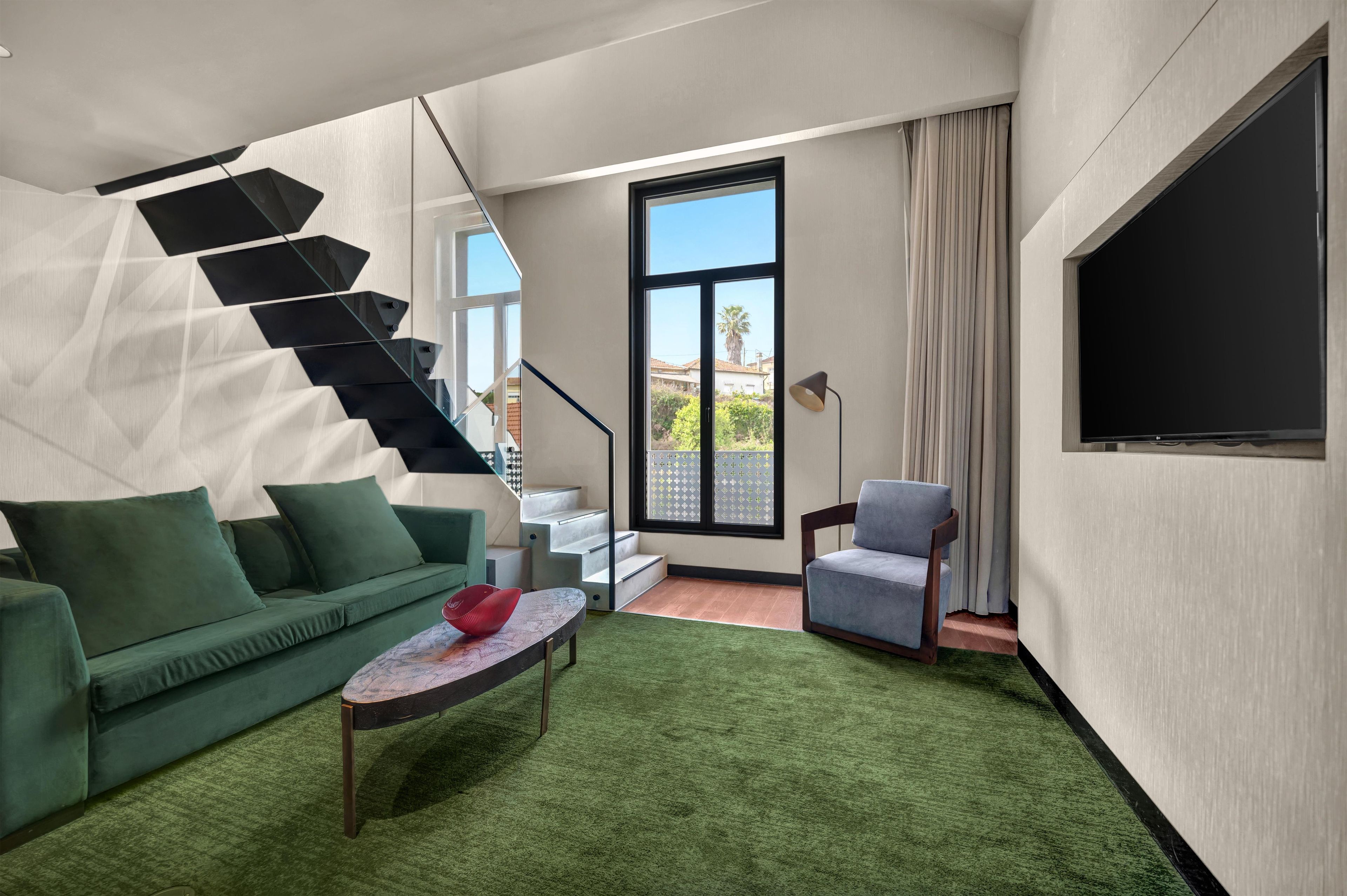 A modern living room with a green sofa, a small table, a blue armchair, a staircase, and a large window with a view of palm trees.