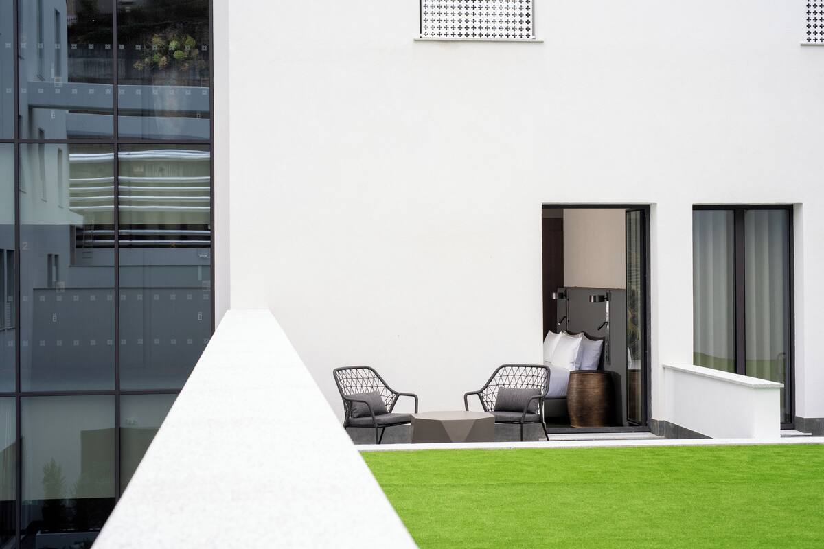 A modern outdoor seating area with two chairs and a small table, a glass wall reflecting the building, and a neatly trimmed lawn.