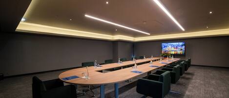 A conference room with a long table, chairs, and a large screen.