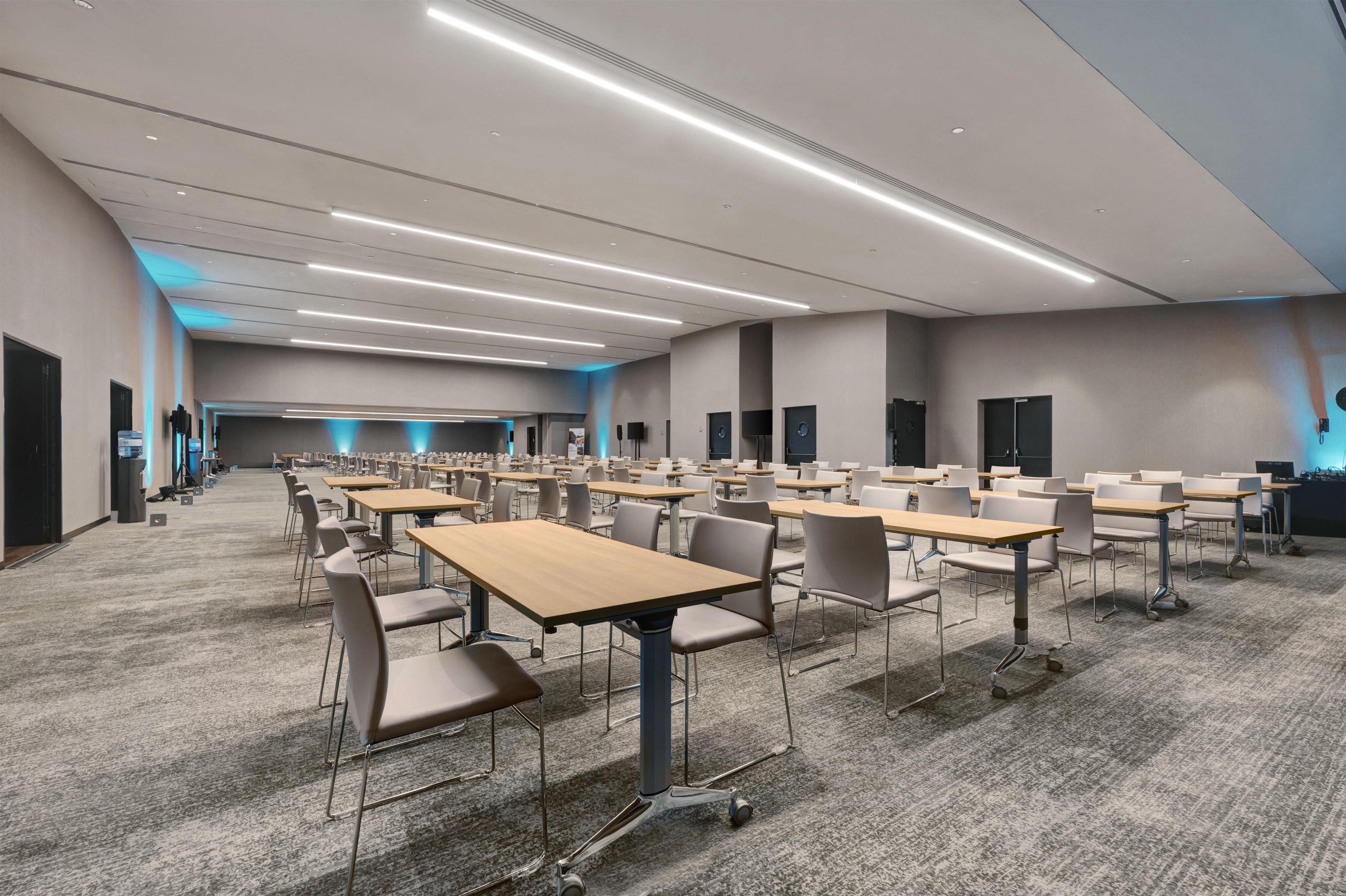 A conference room with rows of tables and chairs, a carpeted floor, and a long ceiling light strip.
