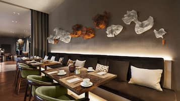A modern dining area with tables set for two, green upholstered chairs, and a dark-colored sofa with cushions.