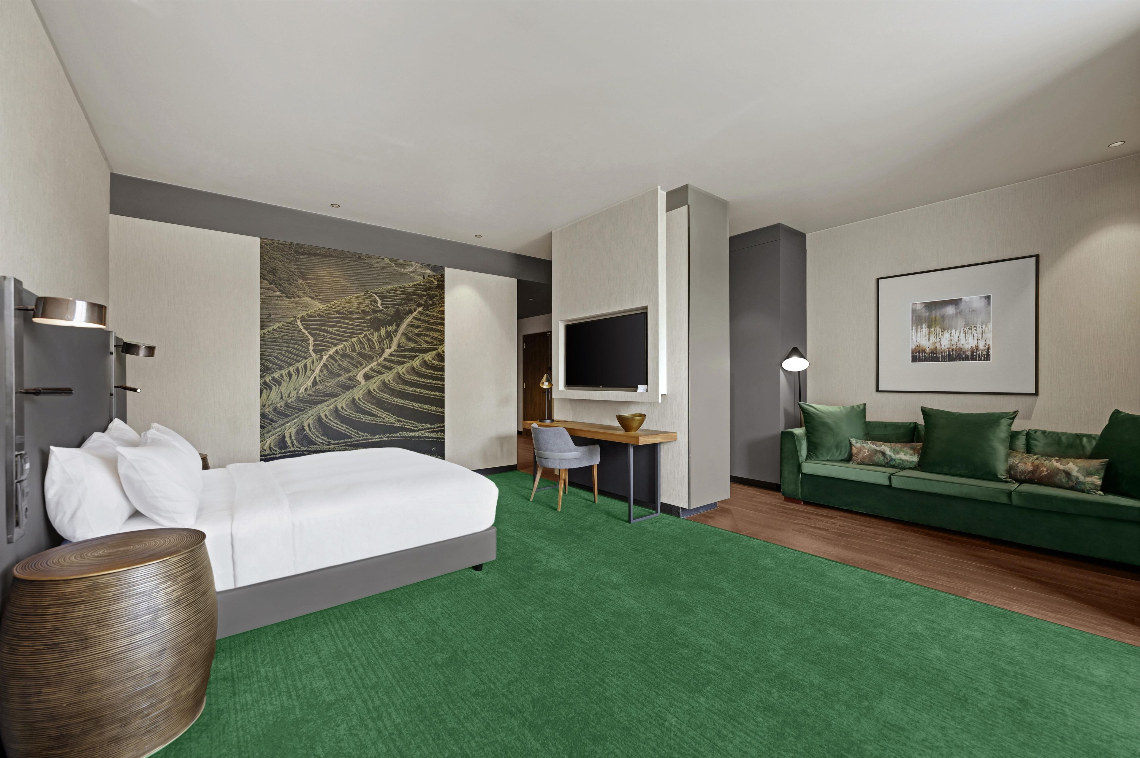 Ett hotellrum med en säng, ett skrivbord, en grön soffa, en tv och ett stort konstverk på väggen.