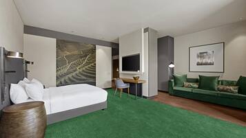Ett hotellrum med en säng, ett skrivbord, en grön soffa, en tv och ett stort konstverk på väggen.