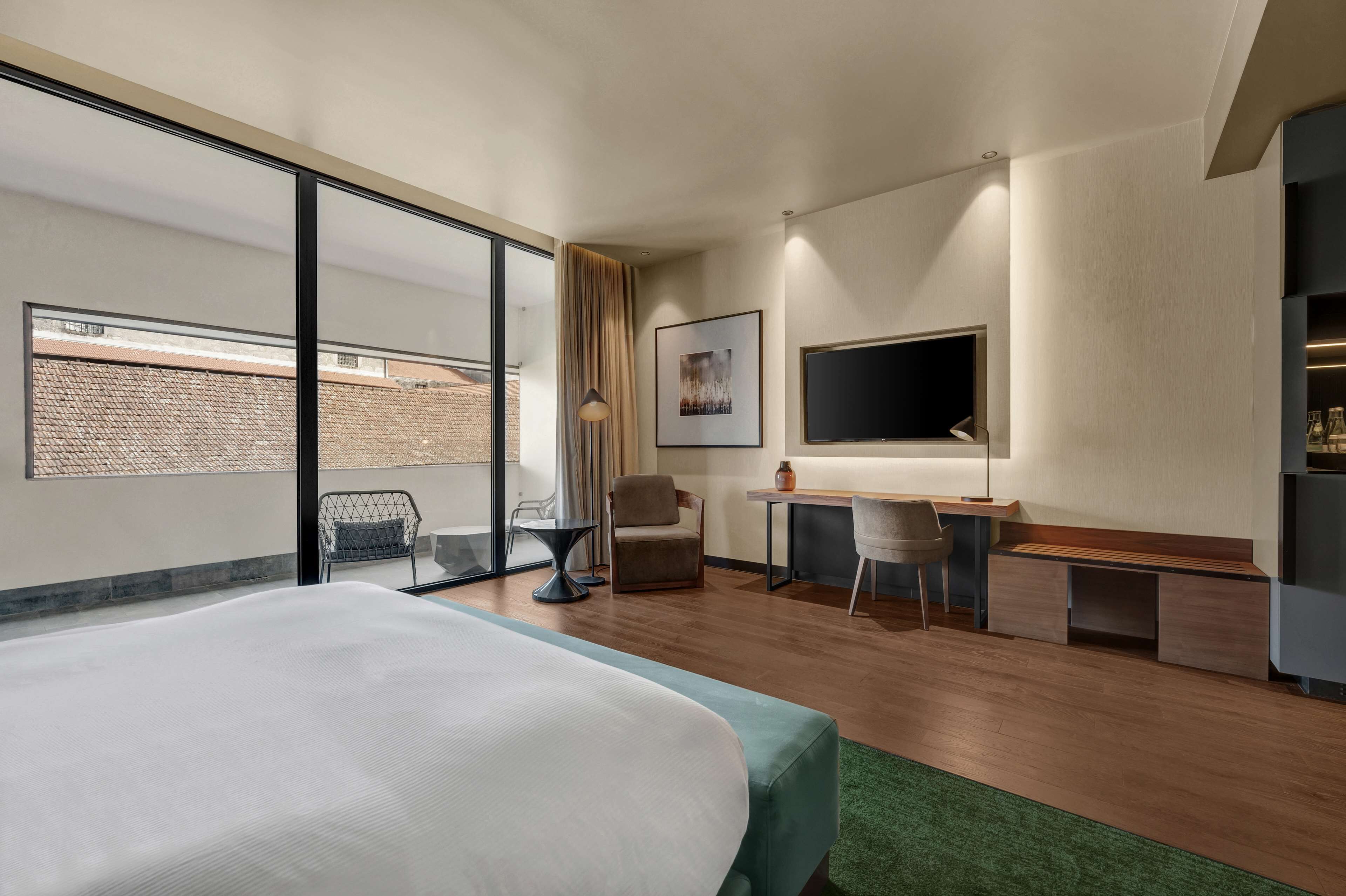 A hotel room with a large bed, a desk, a chair, a TV, and a view of a brick building through a glass door.