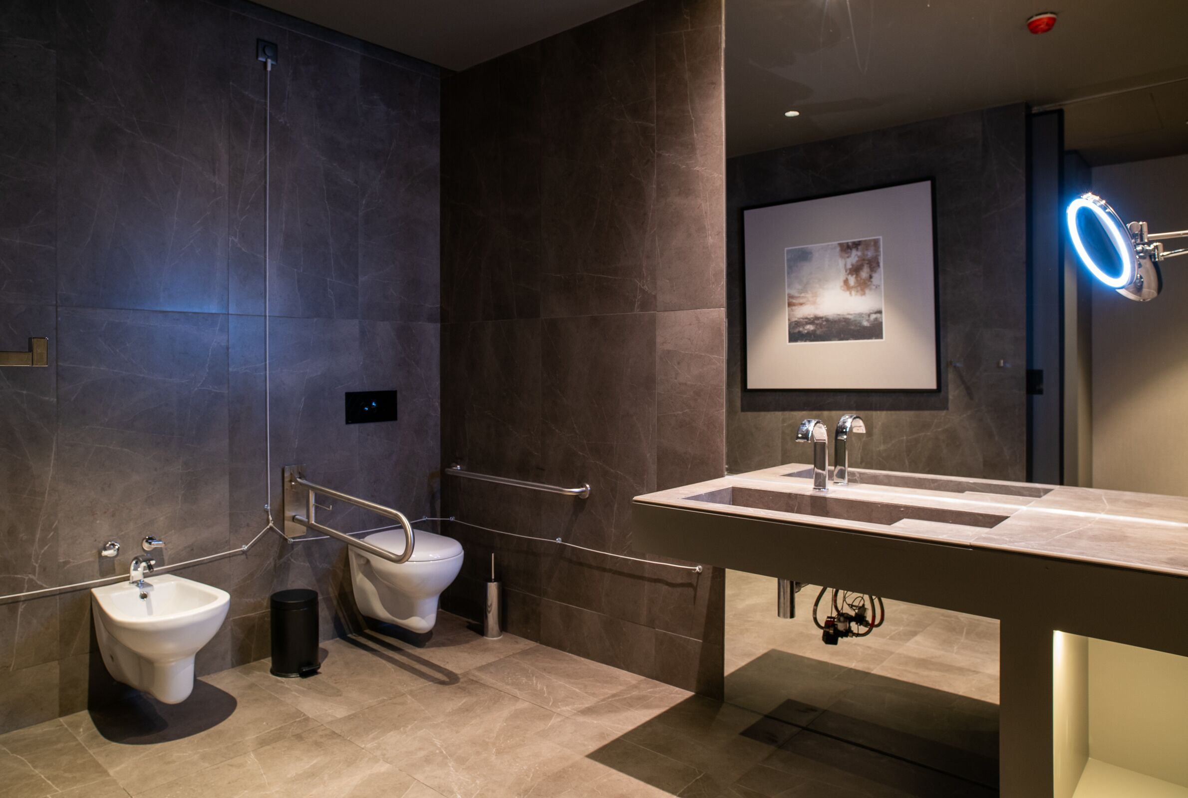 A modern bathroom with a bidet, toilet, vanity with a mirror, and a framed picture on the wall.
