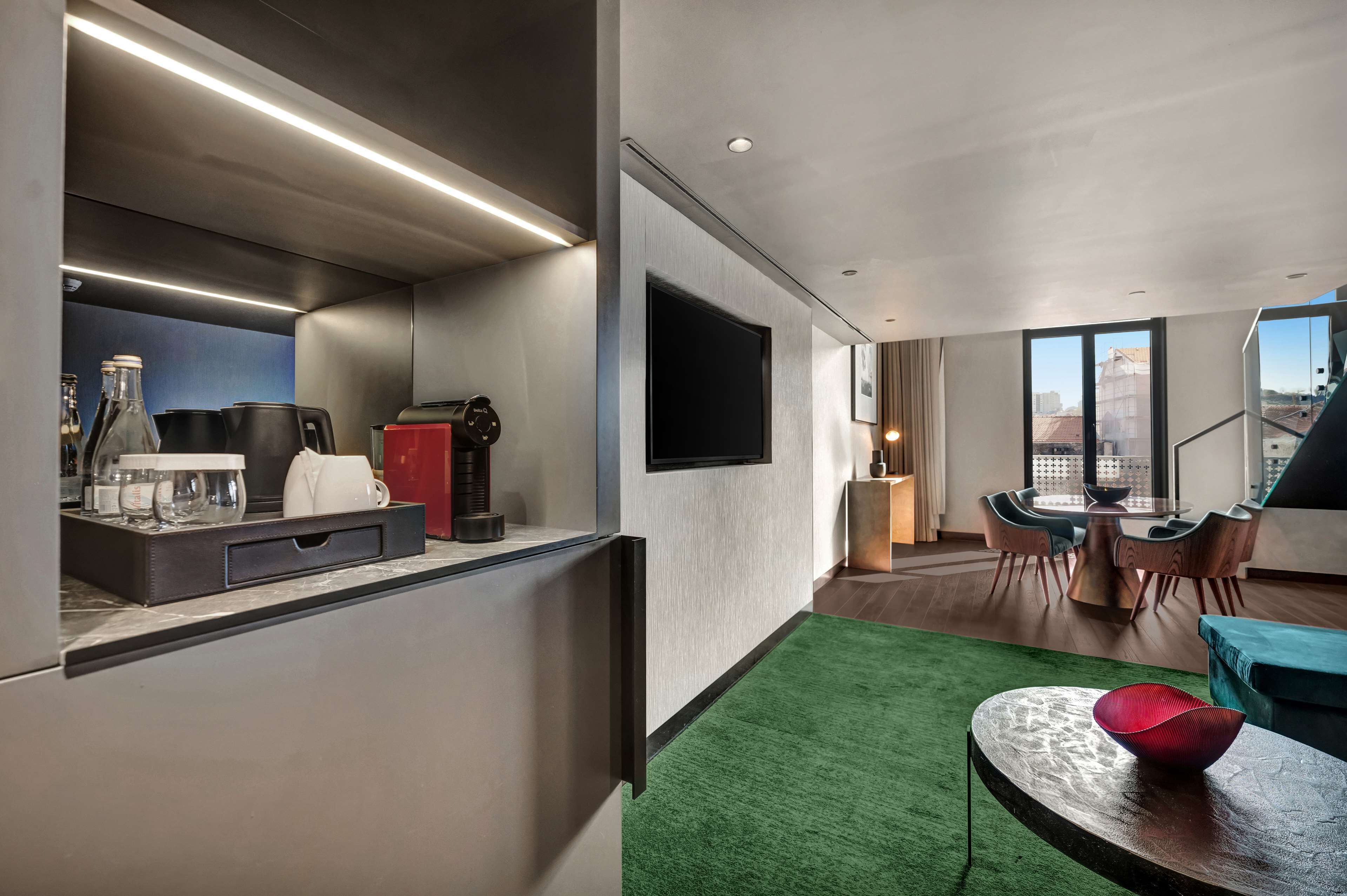 Ein modernes Hotelzimmer mit einer Küchenzeile, einem Flachbildfernseher, einem Essbereich mit Tisch und Stühlen sowie einem Blick auf die Stadt durch große Fenster.
