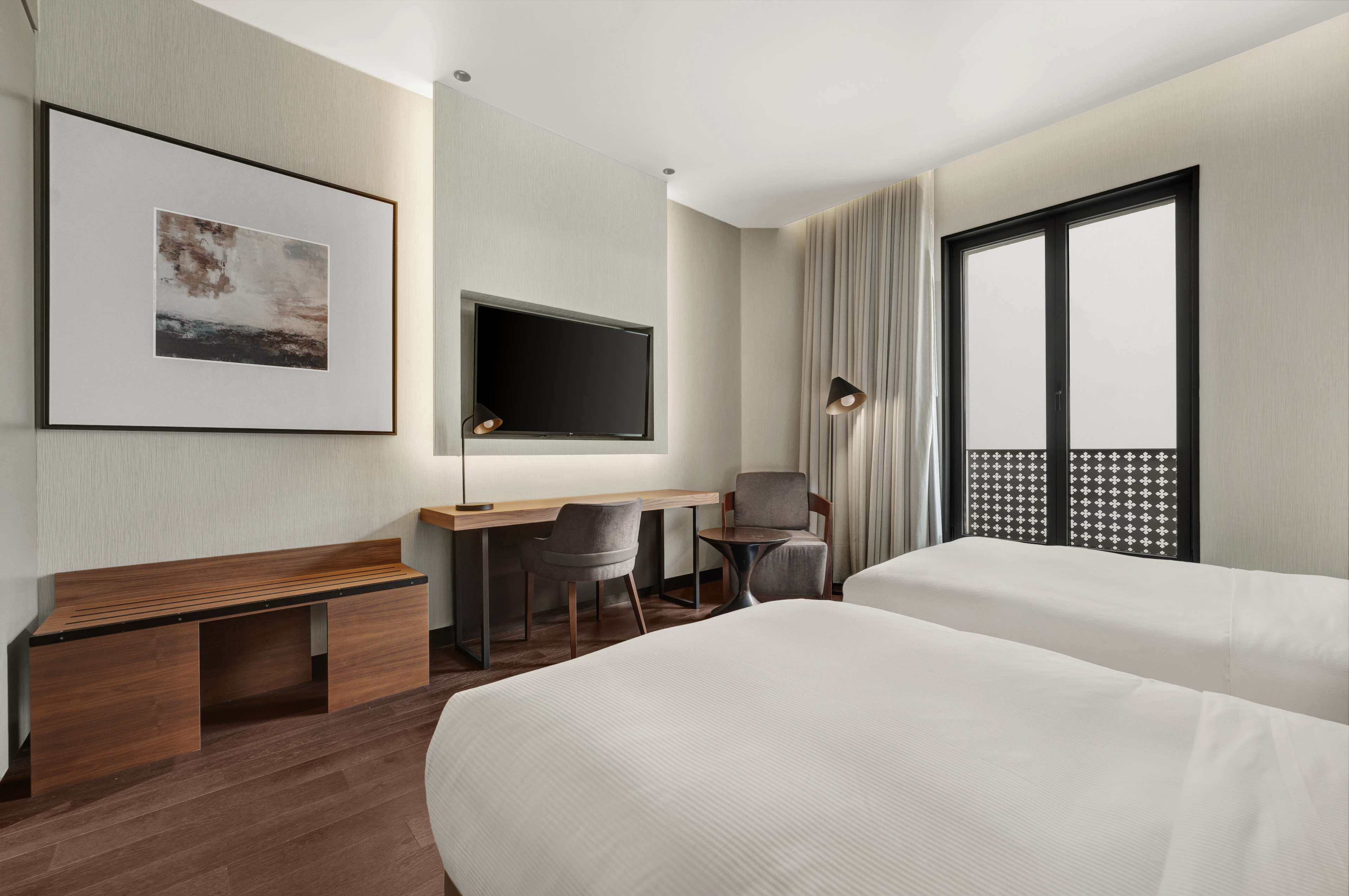 Een hotelkamer met twee bedden, een bureau met stoel, een televisie, een inlijsting foto aan de muur en een groot raam met gordijnen.