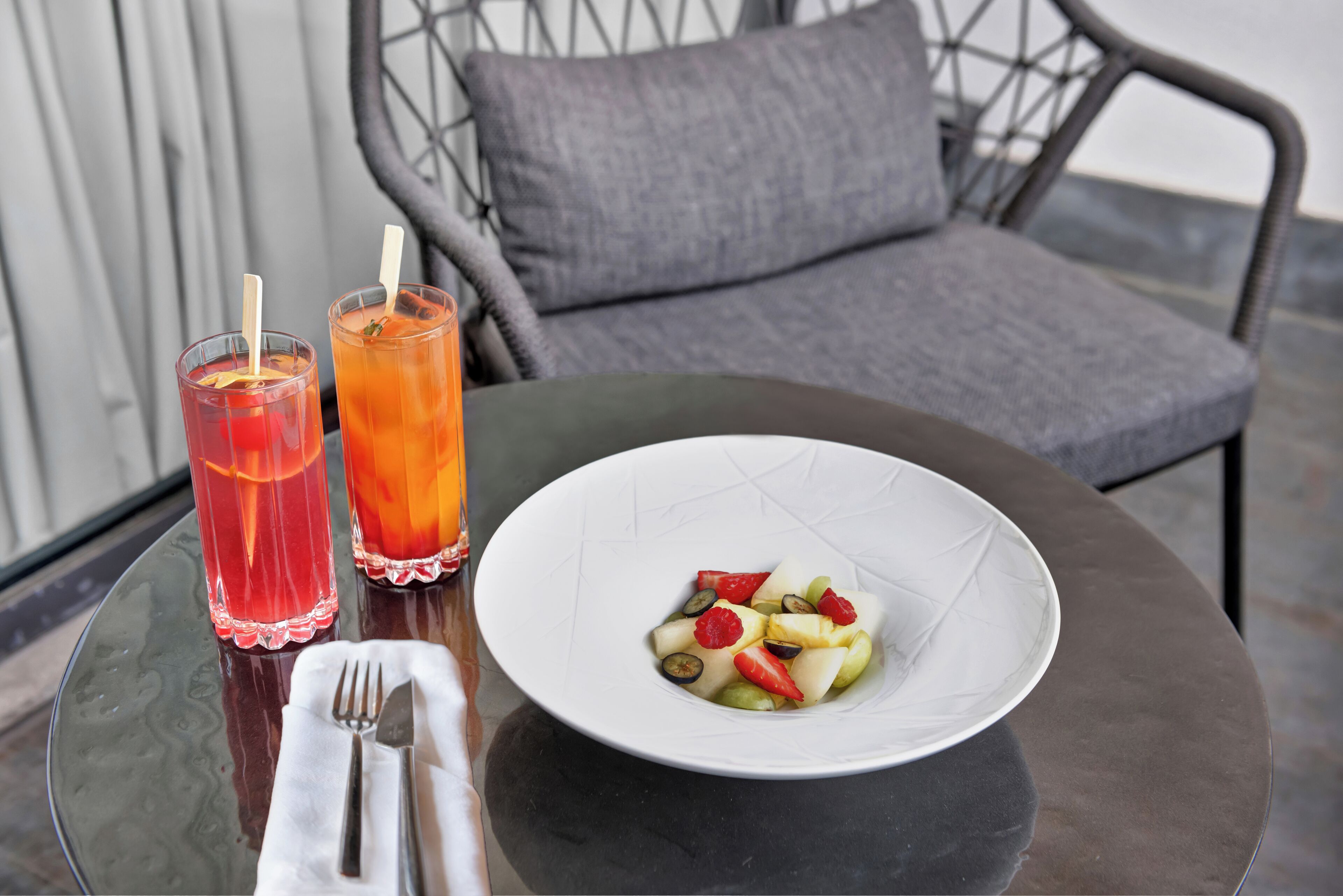 Una mesa con un bol blanco de ensalada de frutas, dos vasos de bebidas heladas y una servilleta doblada con cubiertos sobre una mesa de vidrio.