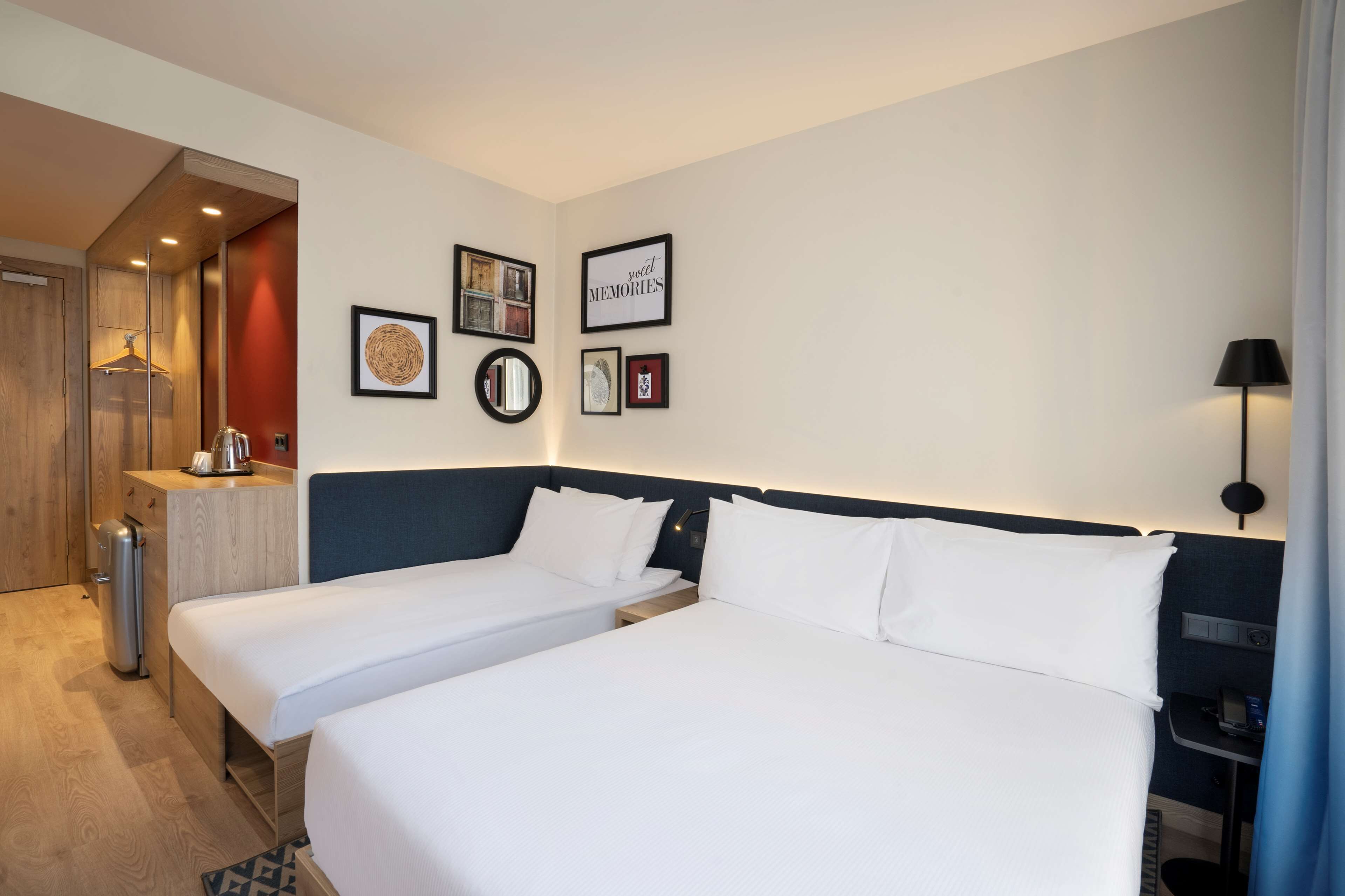 A hotel room with two single beds, a small desk with a lamp, a mirror, and wall art including a 'sweet memories' sign.