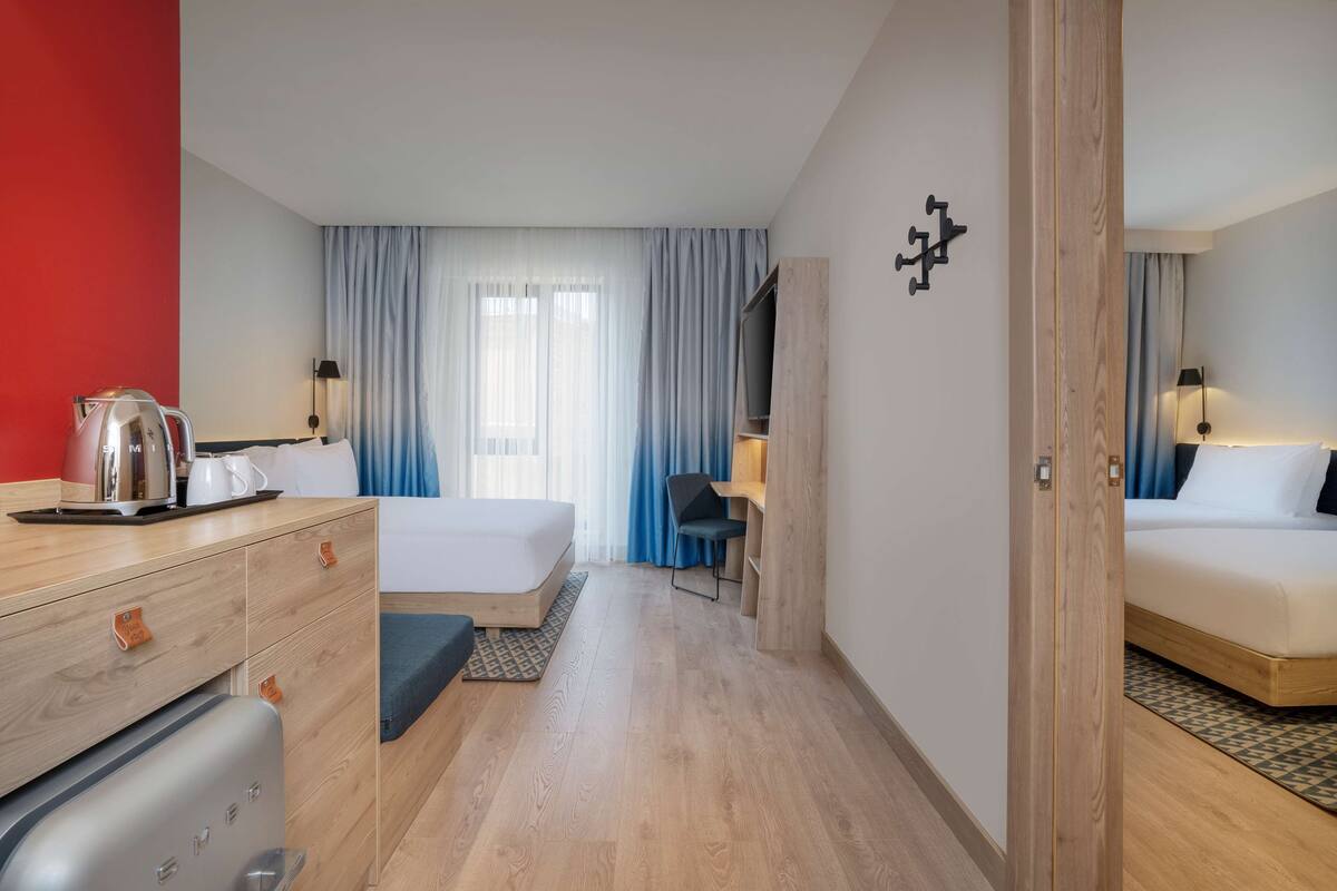 A hotel room with a wooden floor, a bed, a desk with a kettle, a chair, and a TV on a wooden stand.