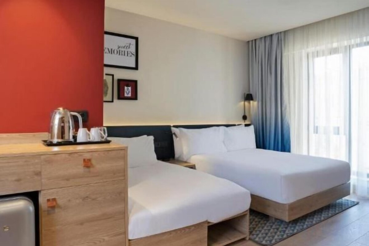 Habitación de hotel con dos camas, un aparador de madera, un escritorio pequeño y una ventana con cortinas.