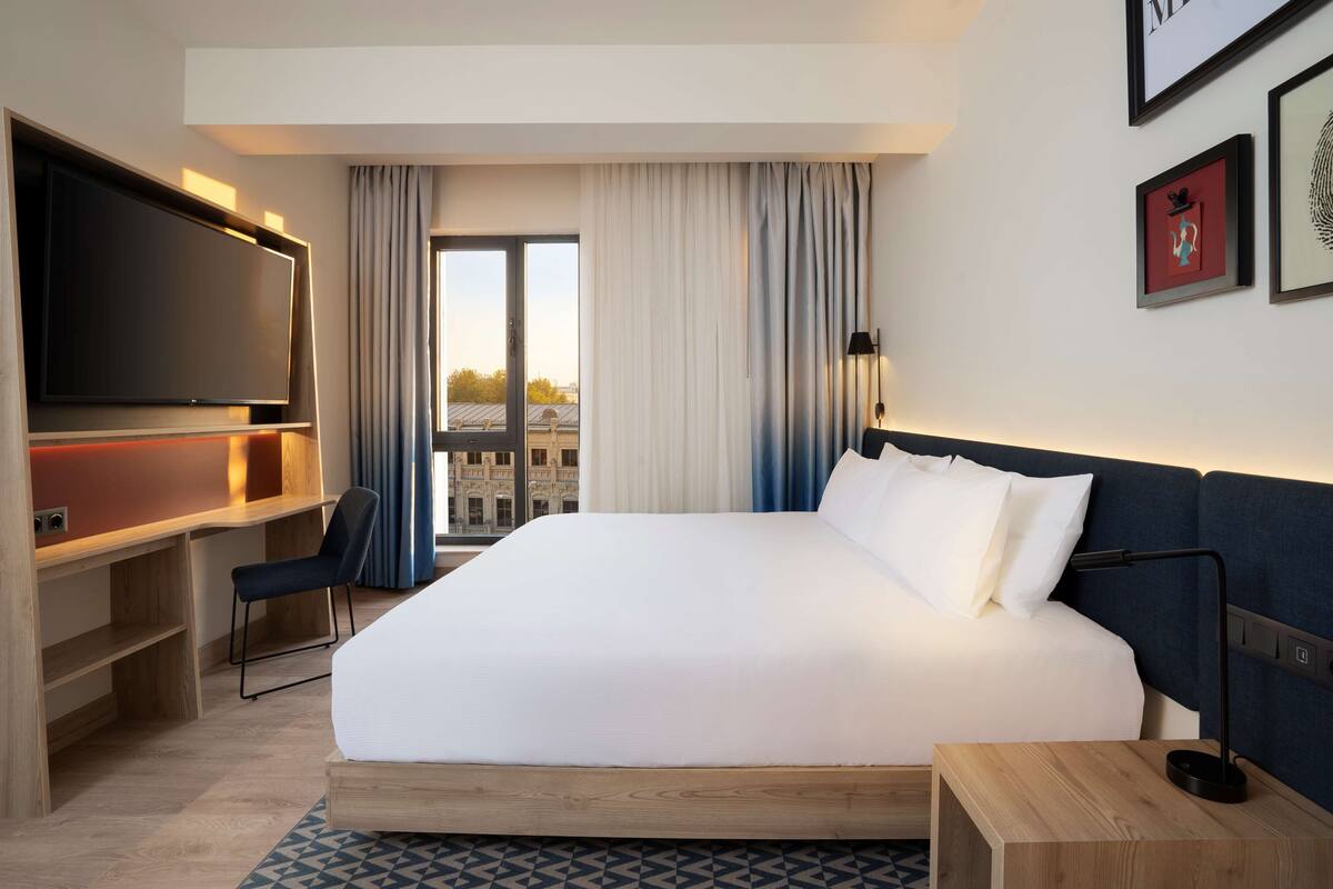 A hotel room with a large bed, a desk with a TV, a chair, and a view of a cityscape through a window.
