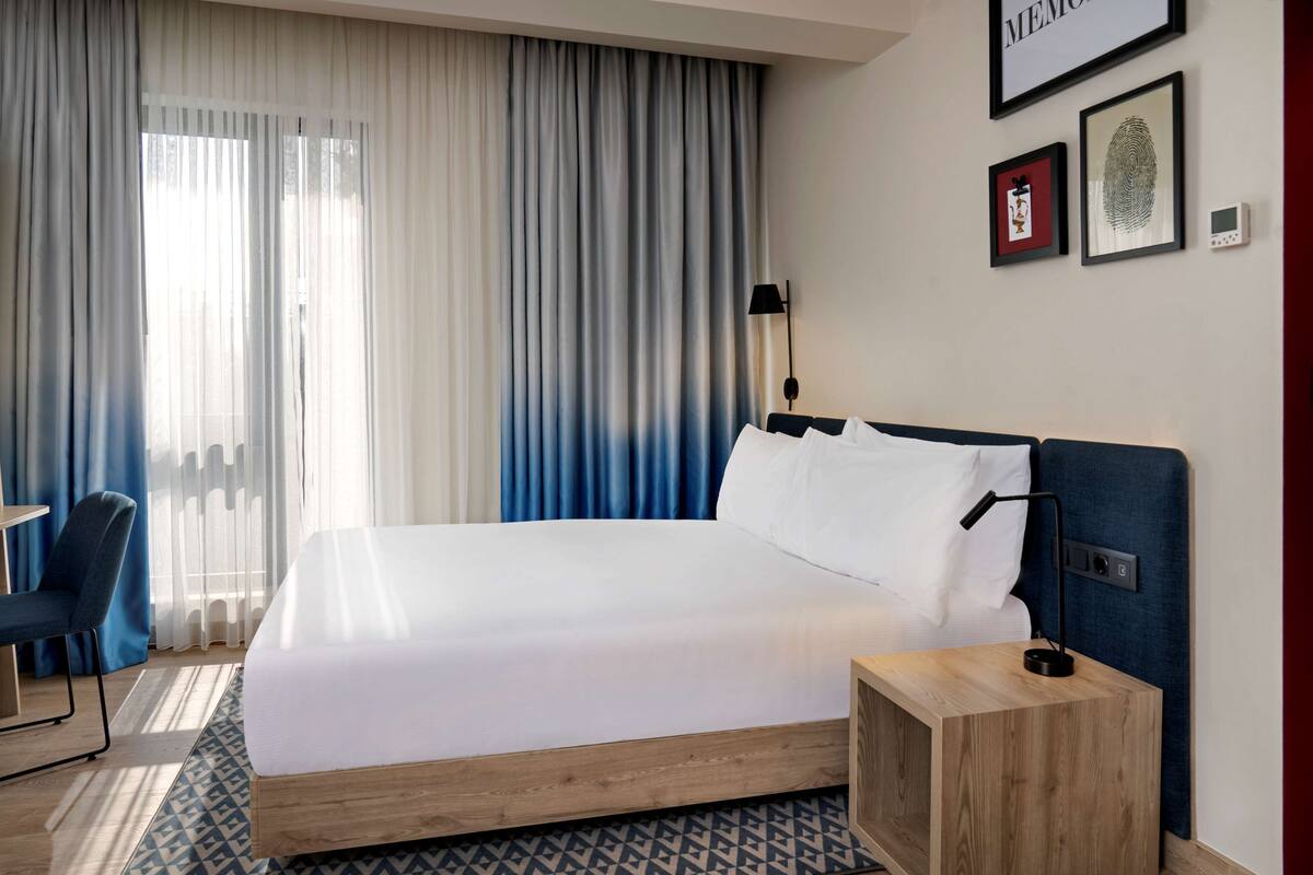A hotel room with a bed, a desk, a chair, a bedside table, and a wall-mounted lamp.
