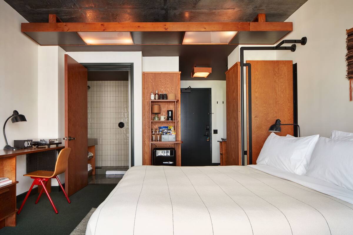 A hotel room with a bed, a desk with a chair, a wardrobe, and a bathroom visible through an open door.