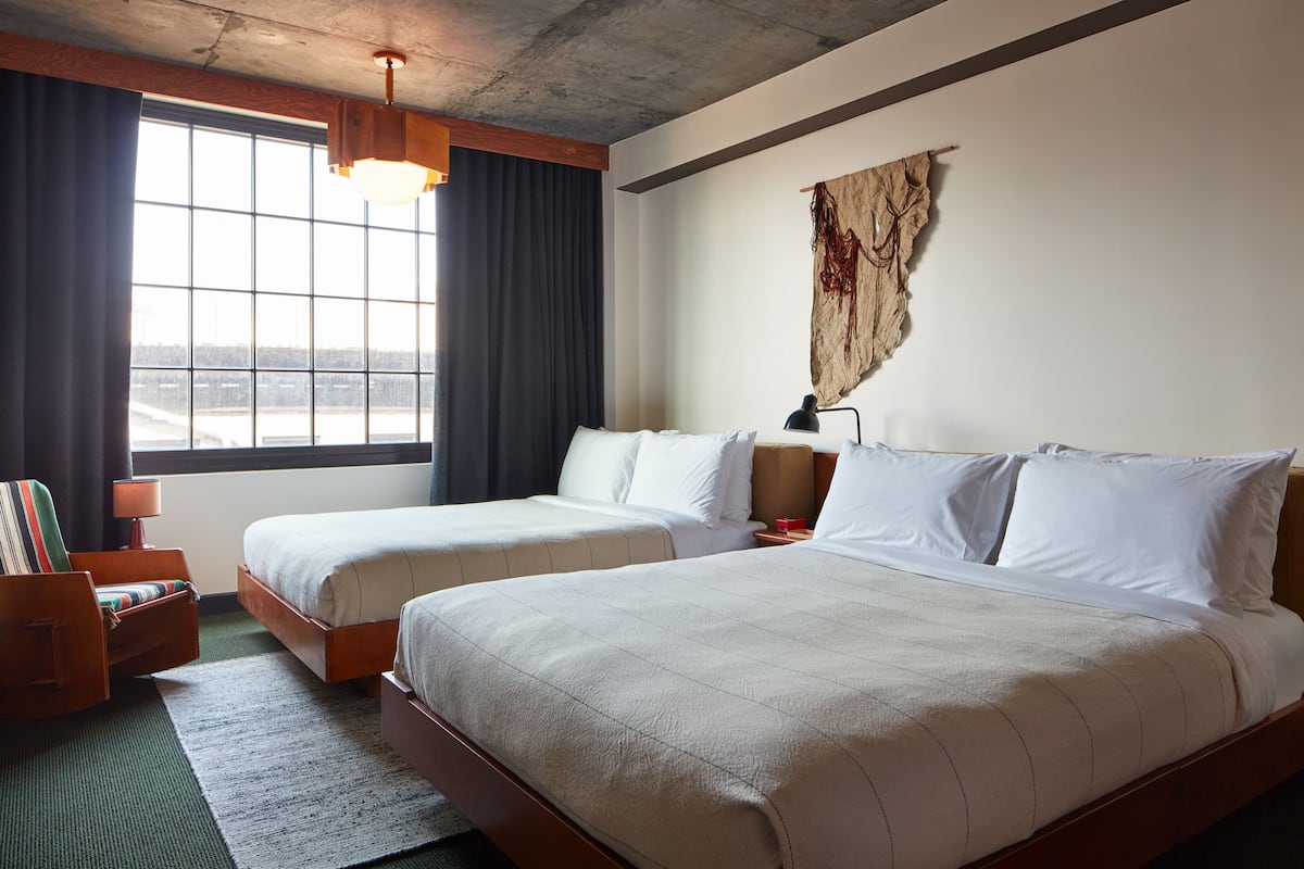 A hotel room with two beds, a large window, a wooden headboard, a wall-mounted tapestry, and a green rug.