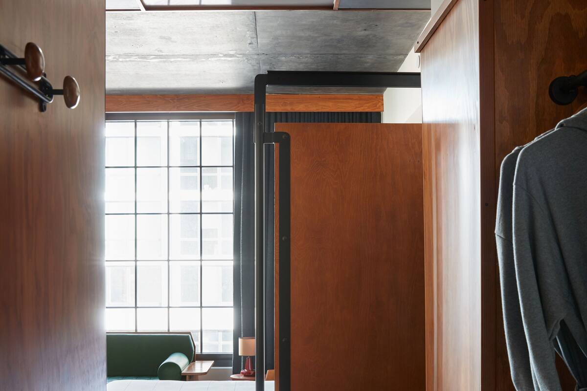 A hotel room with a wooden wardrobe, a bed with a green headboard, a window with blinds, and a green carpet.