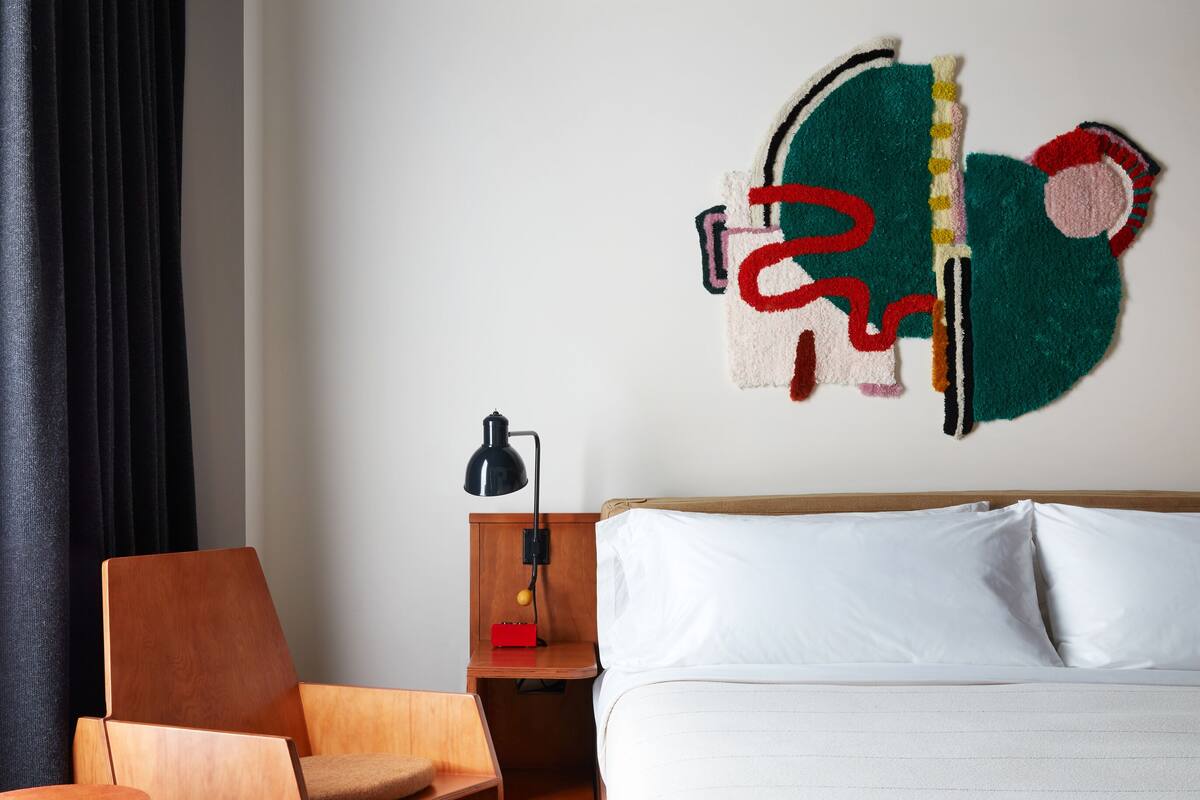 A modern hotel room with a bed, a wooden chair, a nightstand, and a unique abstract wall art piece.