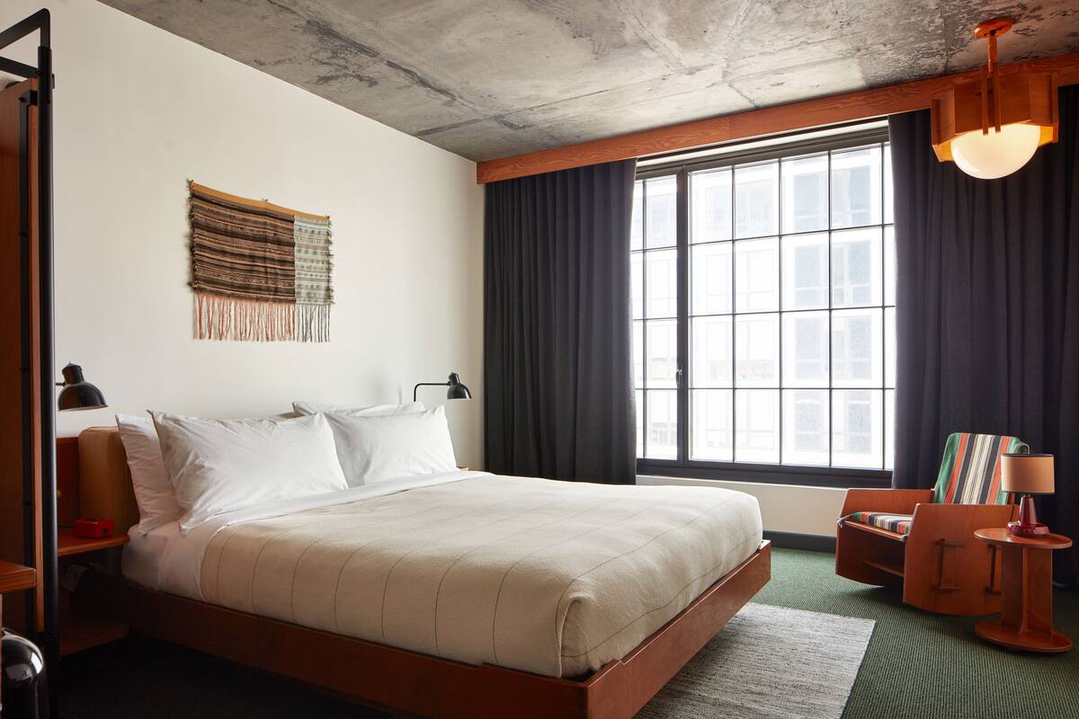 A hotel room with a large bed, a window with curtains, a small table with a lamp, and a wall hanging.