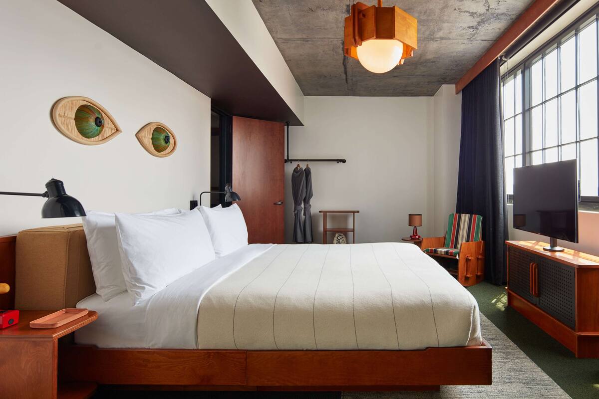 A modern hotel room with a large bed, wooden headboard, a flat-screen TV, and a large window with curtains.