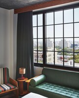 A corner room with a green sofa, striped chair, and a lamp near a window with a city view.