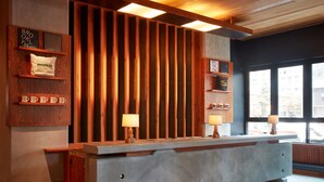 A reception desk with a wooden backdrop, two wall-mounted lamps, and a shelf with branded mugs. - Ace Hotel Brooklyn (Brooklyn)
