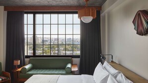 A hotel room with a large bed, a green sofa, a view of the city through a large window, and a lamp on a bedside table. - Ace Hotel Brooklyn (Brooklyn)