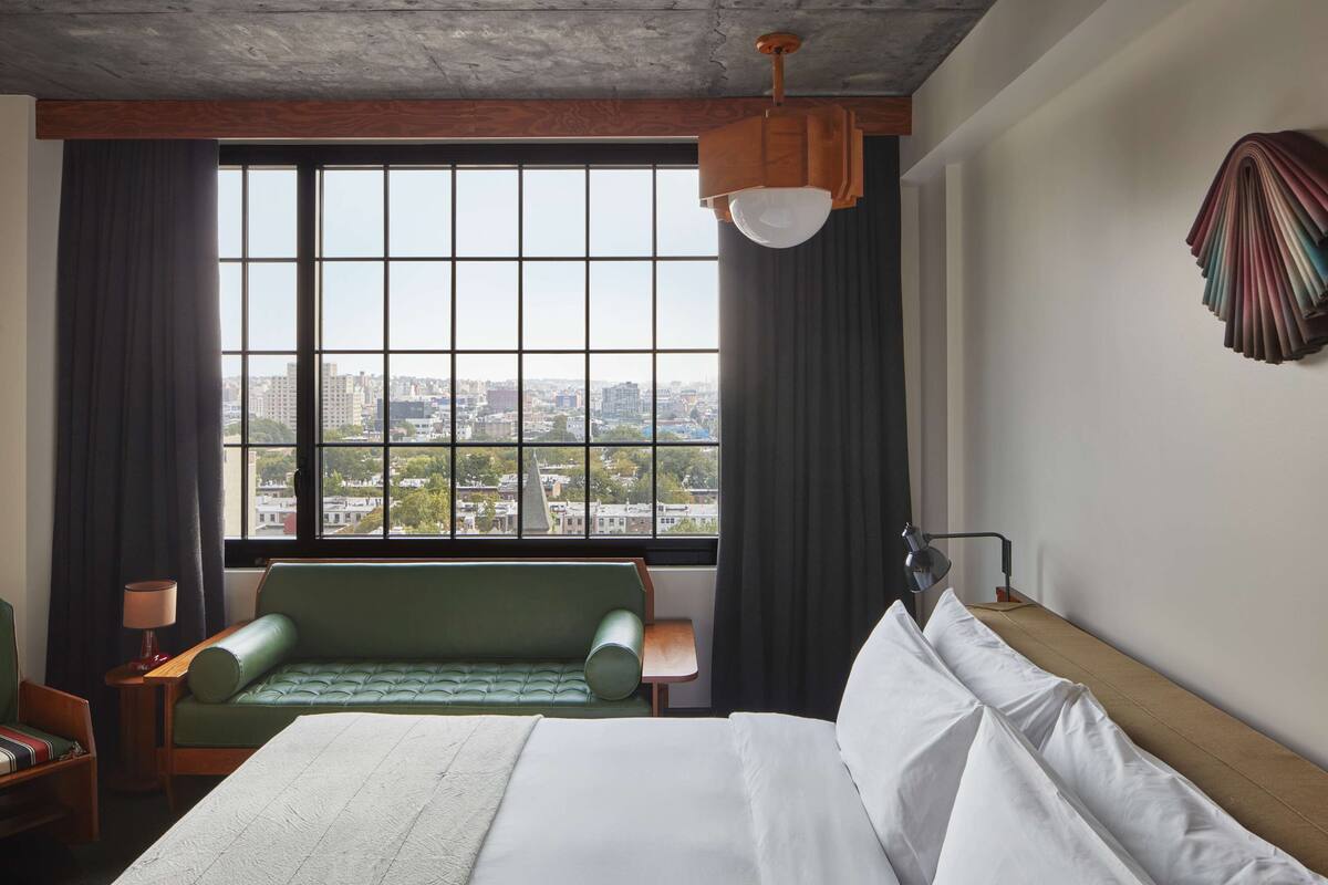 A hotel room with a large bed, a green sofa, a view of the city through a large window, and a lamp on a bedside table.