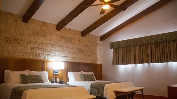 A room with two beds, wooden beams on the ceiling, a stone wall, a window with curtains, and a bedside table with a lamp. - Los Brezos Boutique Hotel (Volcan)
