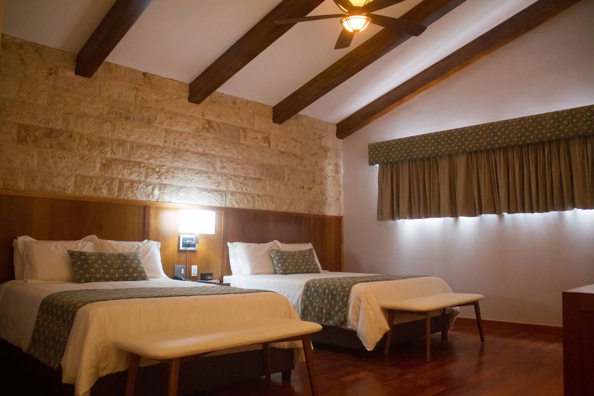 A room with two beds, wooden beams on the ceiling, a stone wall, a window with curtains, and a bedside table with a lamp.